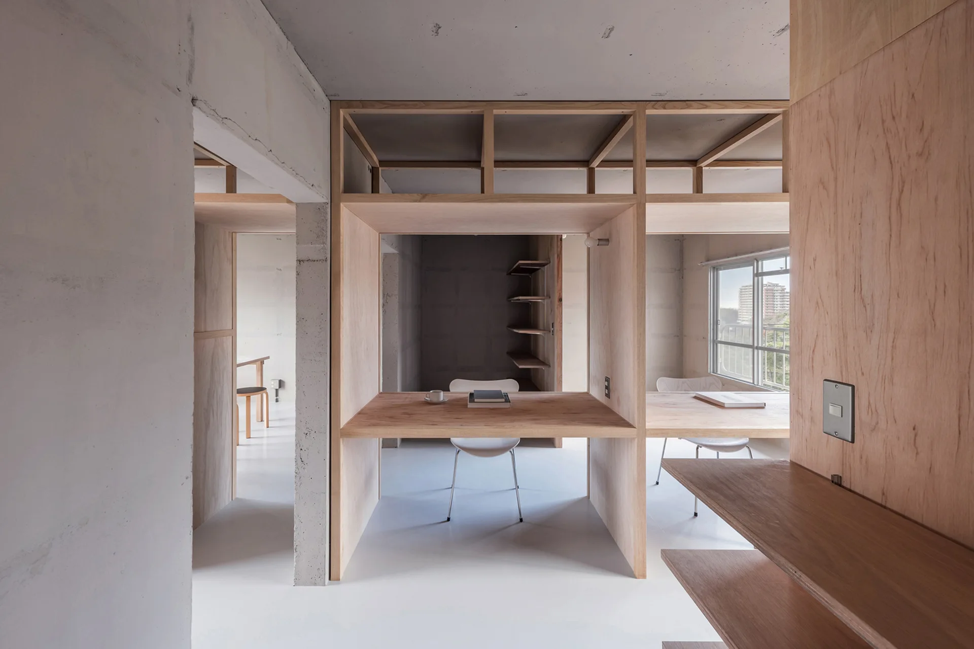 A minimalist workspace featuring a plywood desk partition structure, exposed concrete walls, and white flooring.
