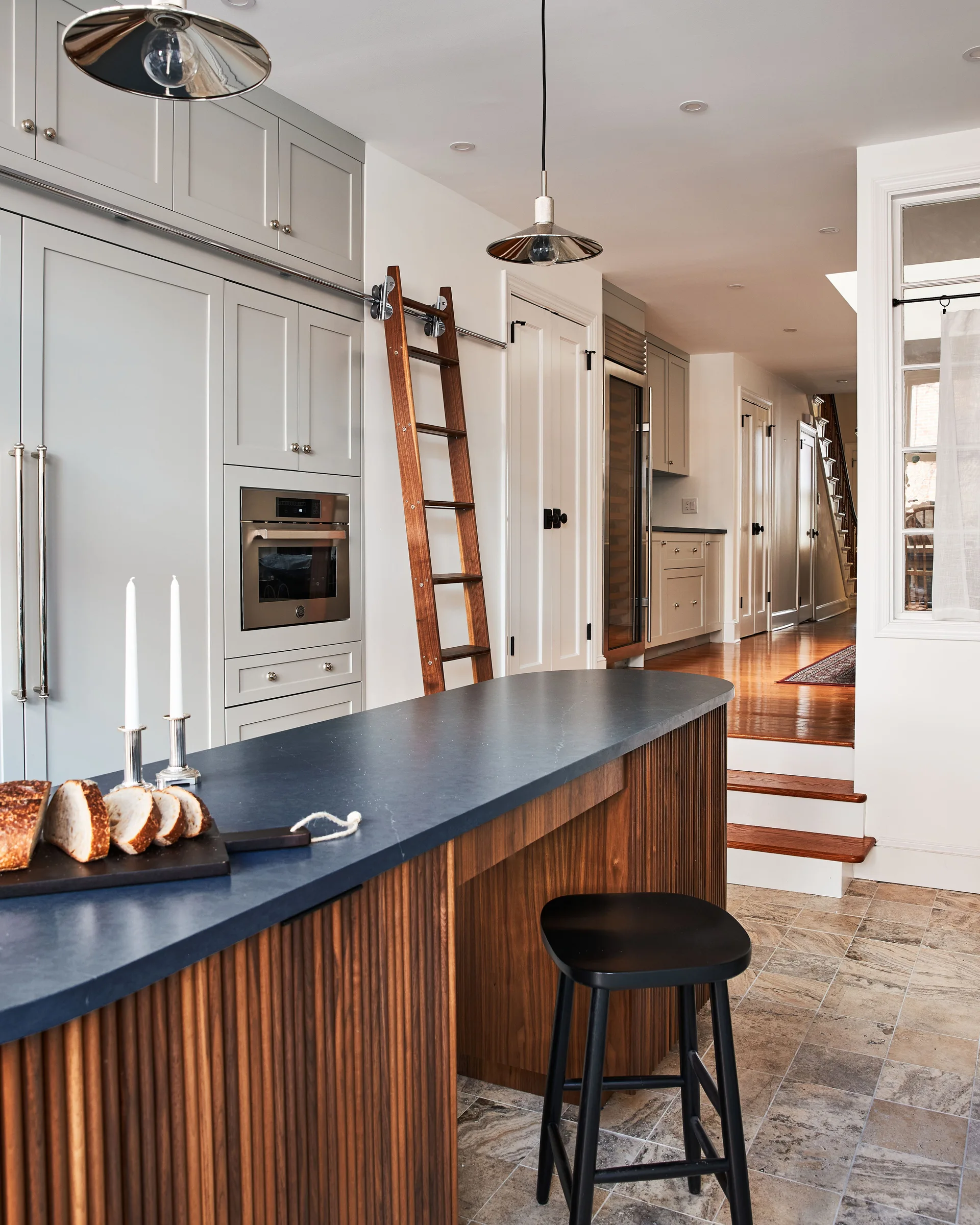 A modern kitchen featuring a fluted wood island with a dark countertop, a black stool, grey cabinets, and a wooden rolling ladder.