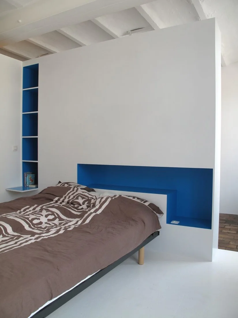 A low bed sits against a white partition wall featuring a long recessed niche and built-in vertical shelving lined in bright blue.