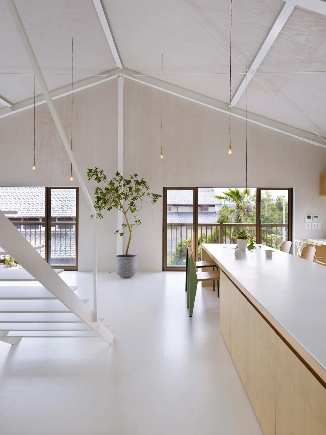 A minimalist open-plan kitchen featuring a long wooden island, a white metal staircase, and light plywood walls under a vaulted ceiling.