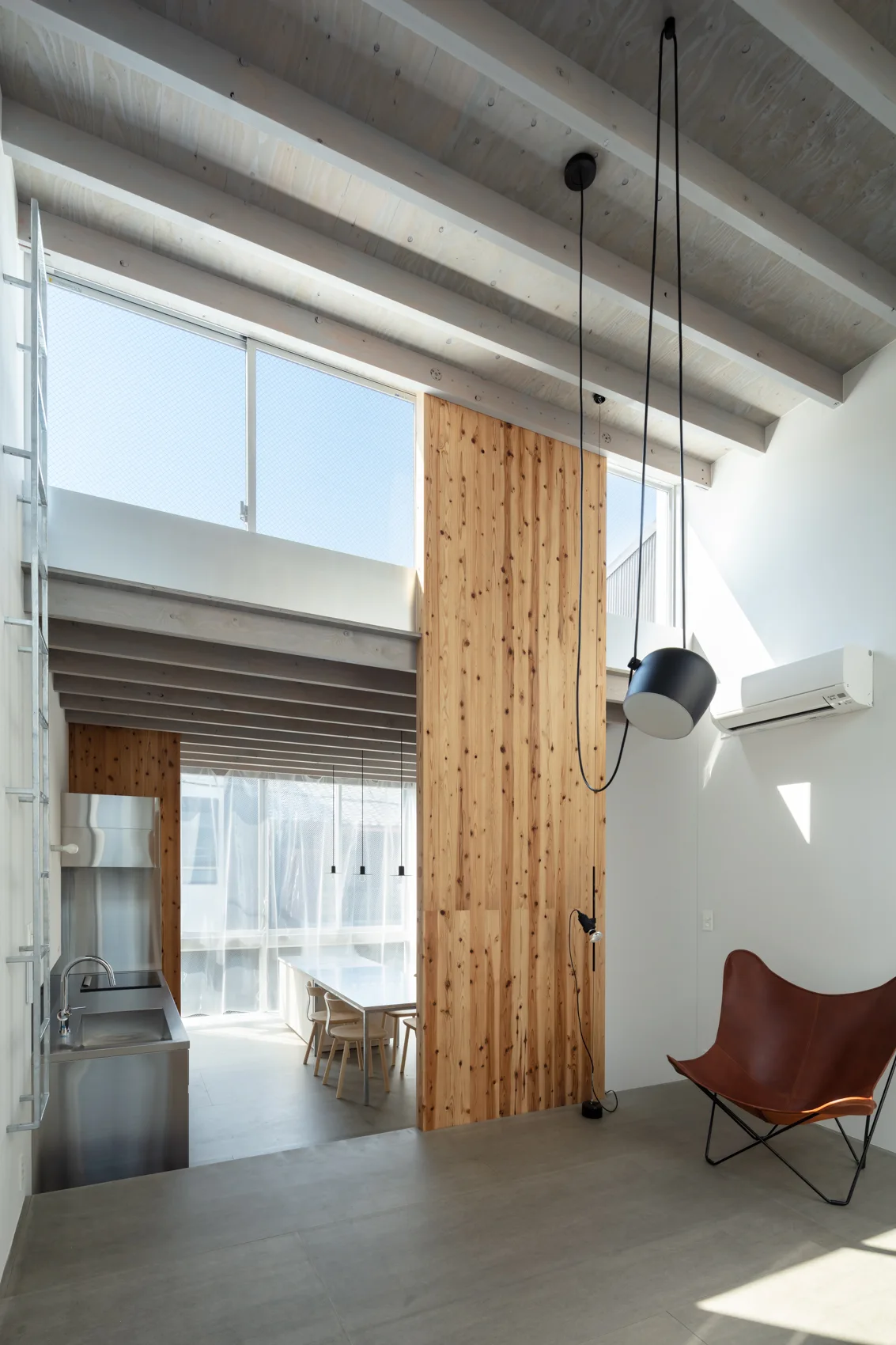 A modern loft interior with exposed wooden ceiling beams, a vertical pine accent wall, a brown leather butterfly chair, and a stainless steel kitchen.