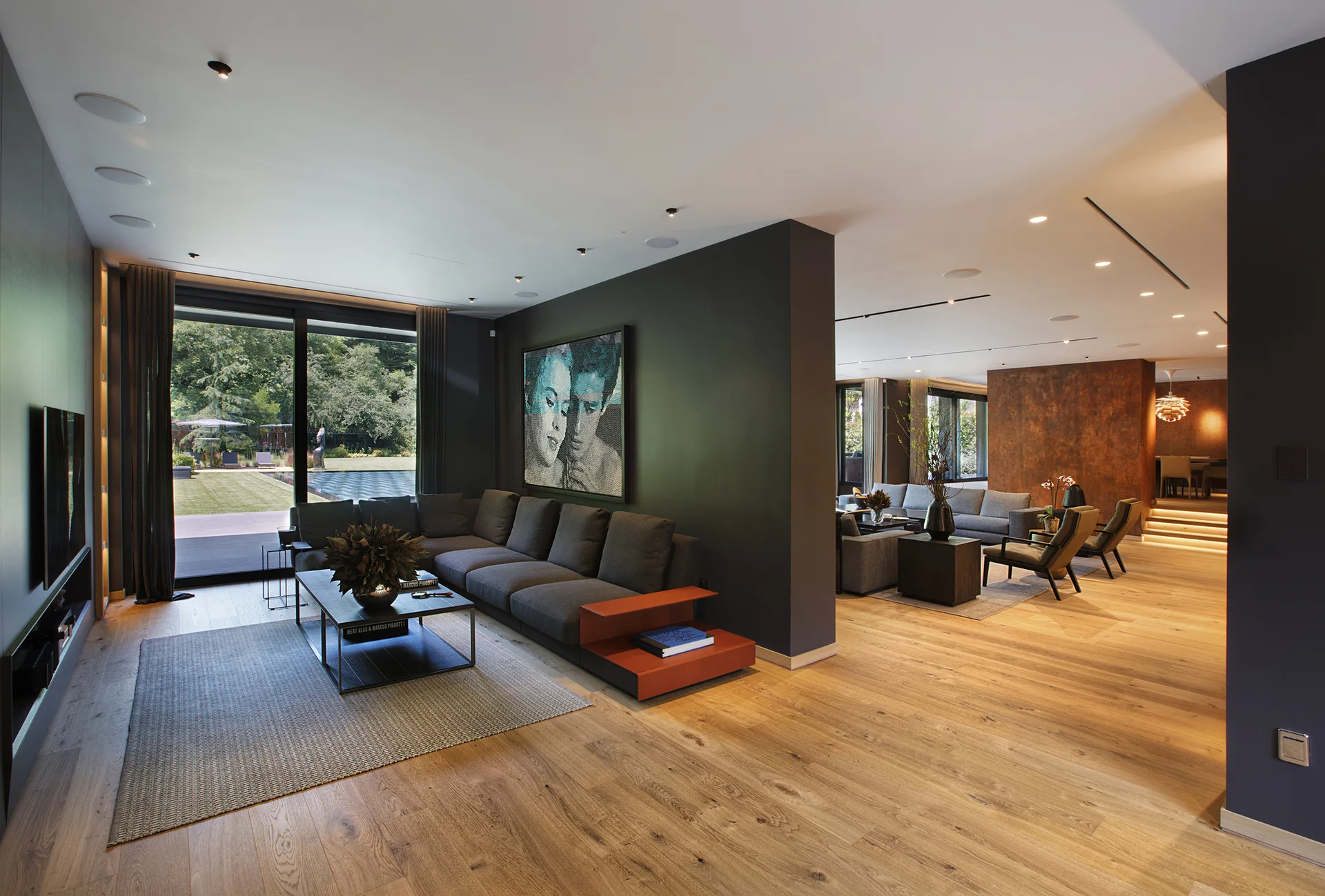 A spacious modern living room features a charcoal grey sectional sofa, a dark central divider wall, and light oak hardwood floors.