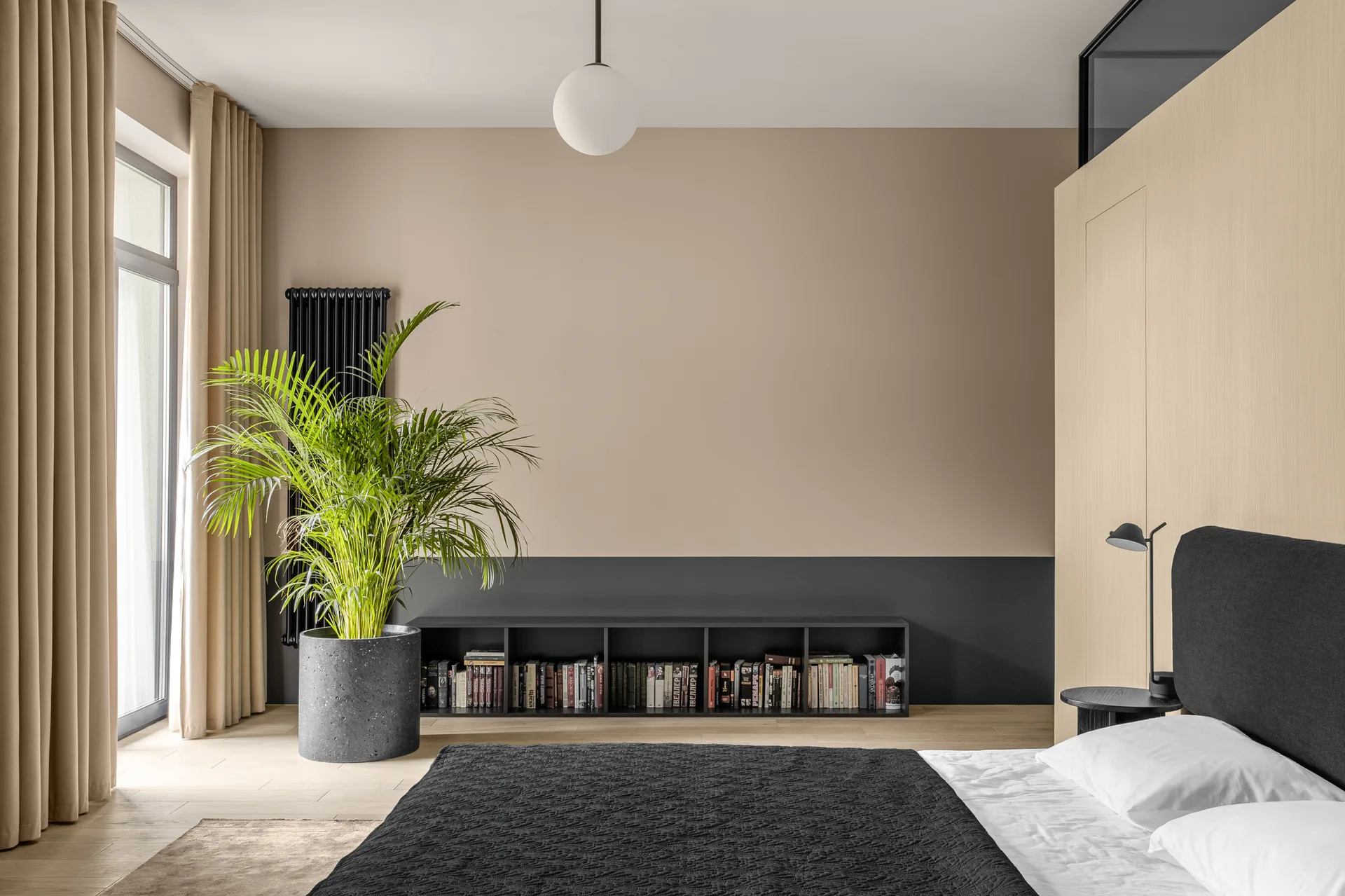 A modern bedroom featuring a two-toned beige and black wall behind a low black bookshelf, a large potted plant, and a bed with a dark headboard.