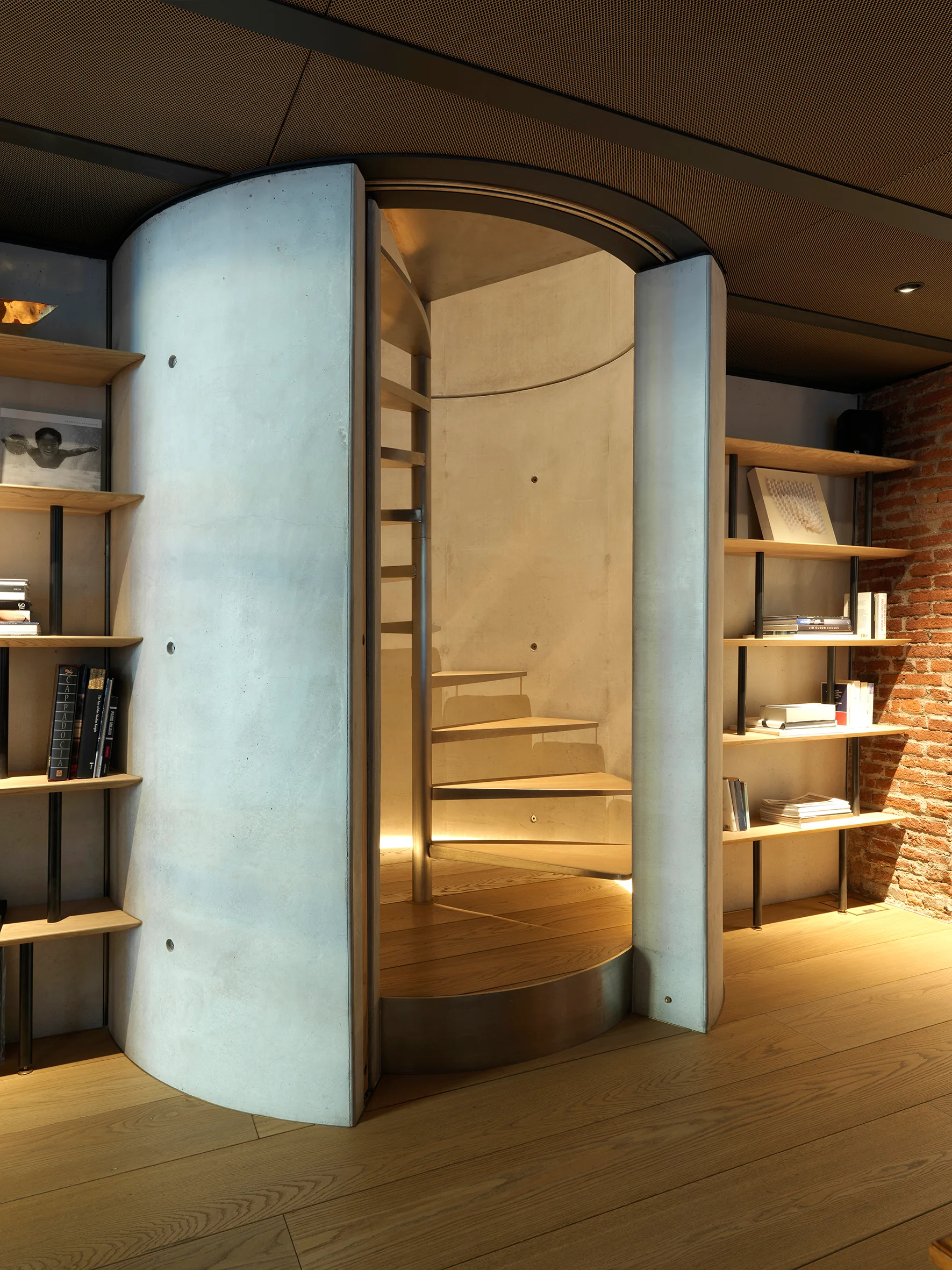 A modern spiral staircase enclosed in a smooth curved concrete column, flanked by open wooden bookshelves against a brick wall on wood floors.