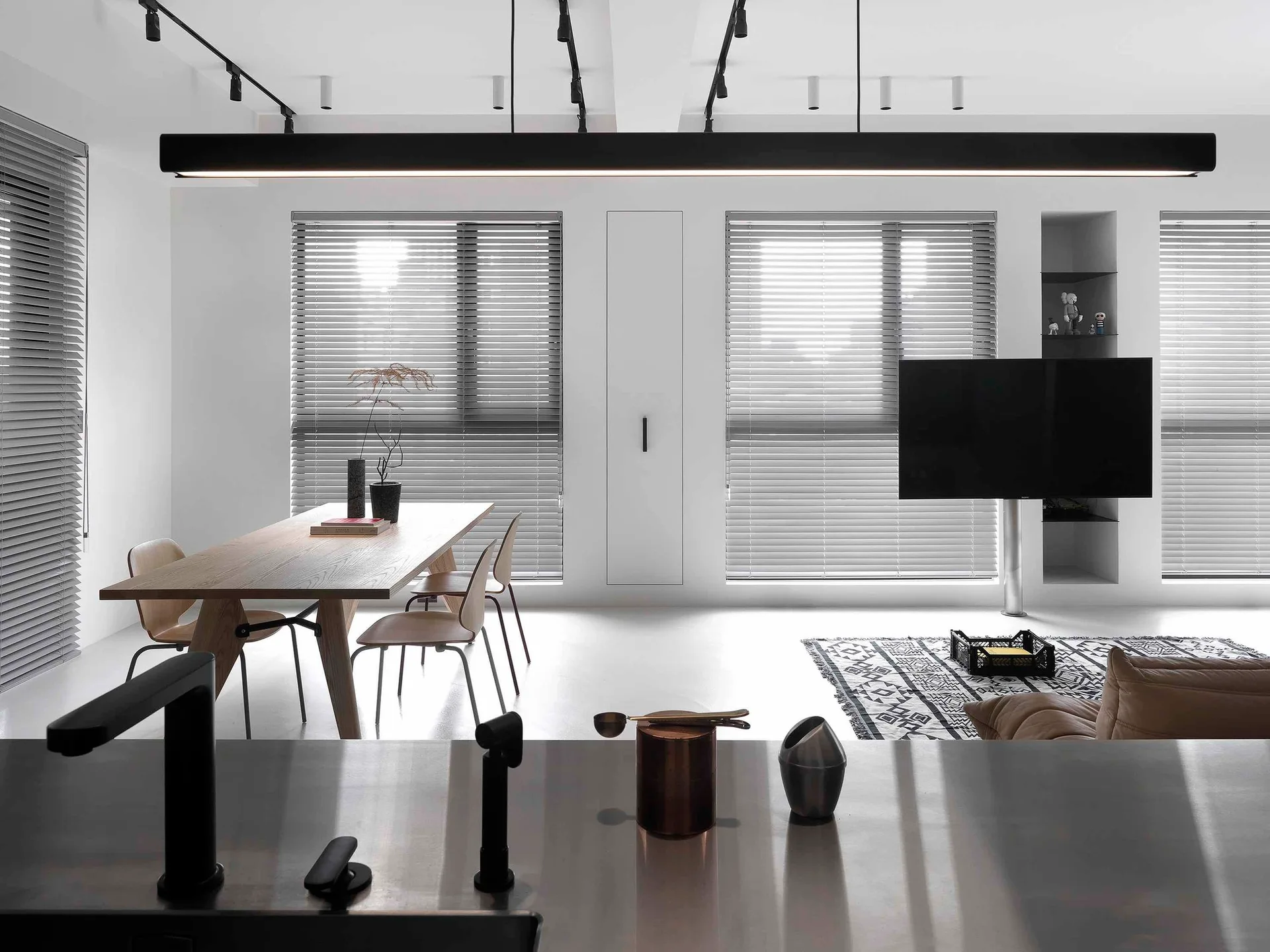 A minimalist dining and living area featuring a wooden table, matching chairs, black track lighting, and grey venetian blinds.