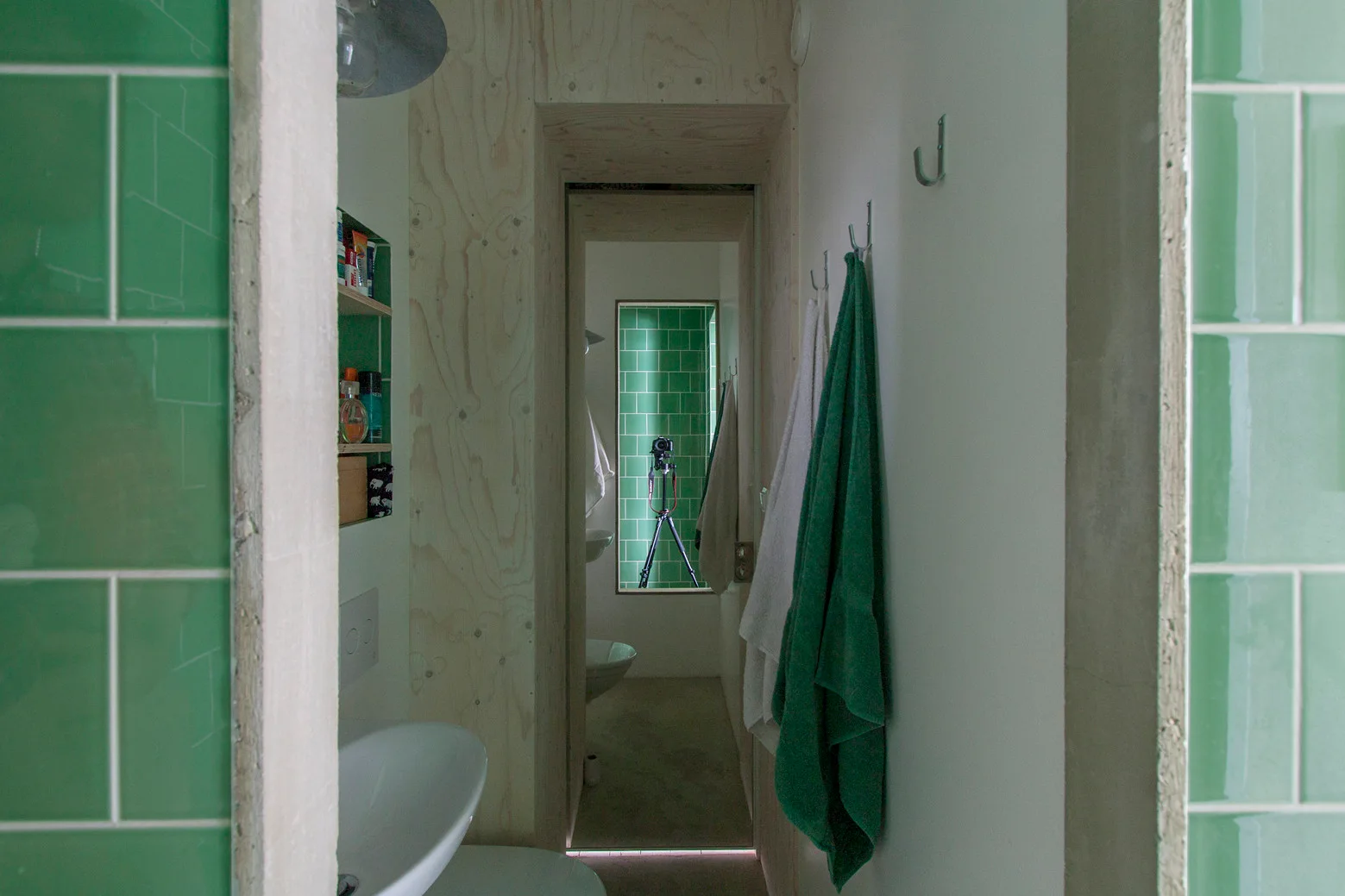 A narrow bathroom corridor features bright green tiled walls, light plywood paneling, and white walls with hanging towels.