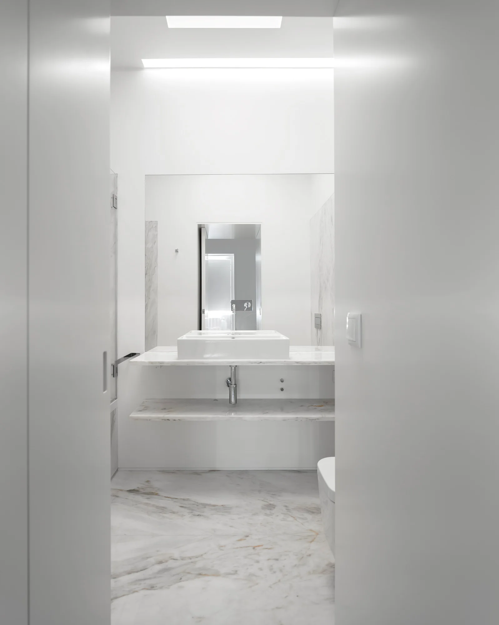 Looking through a doorway into a minimalist white bathroom with a floating marble vanity, rectangular vessel sink, and veined marble flooring.