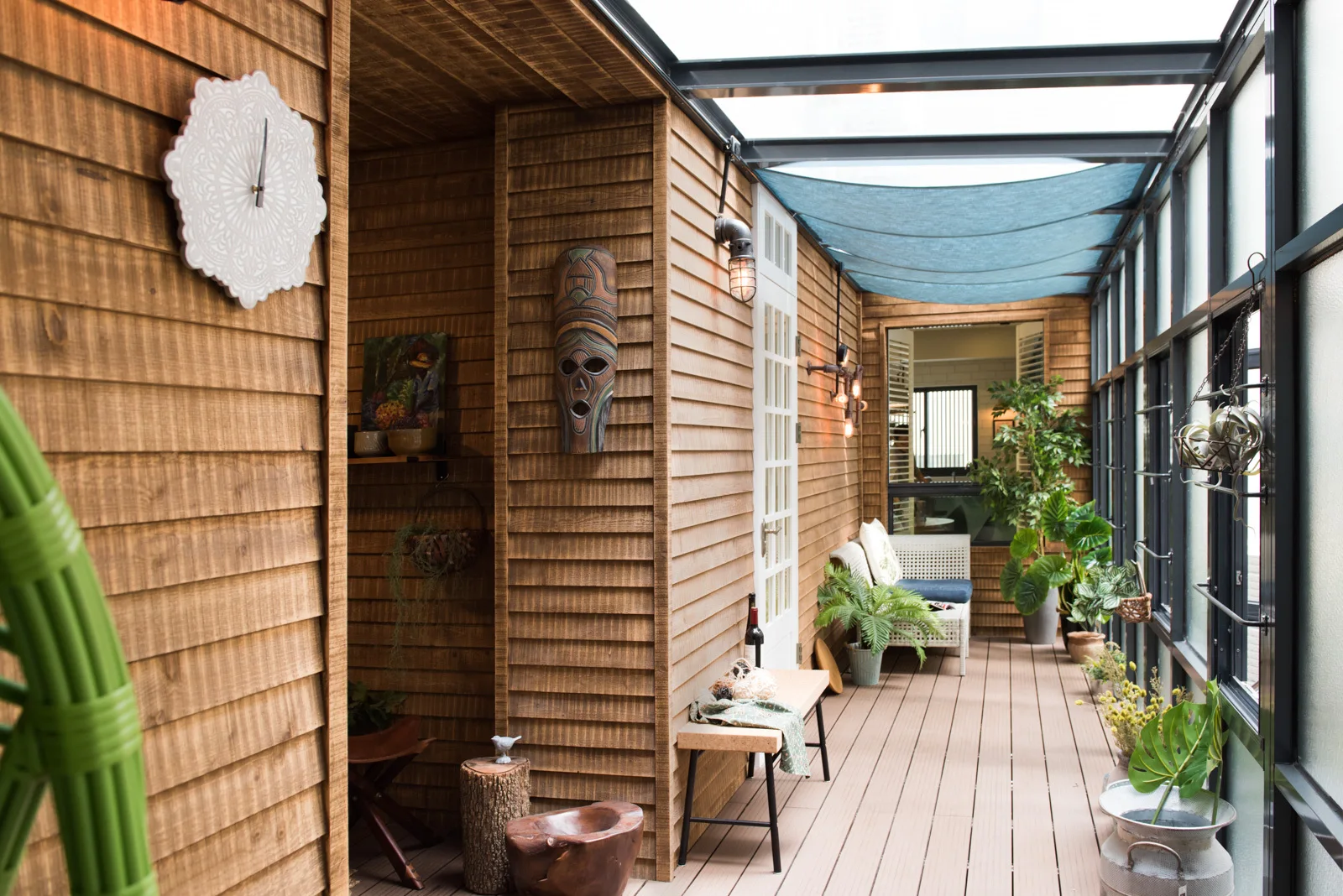An enclosed patio featuring horizontal wood siding, a glass ceiling with a blue shade canopy, and potted plants alongside a wooden deck floor.