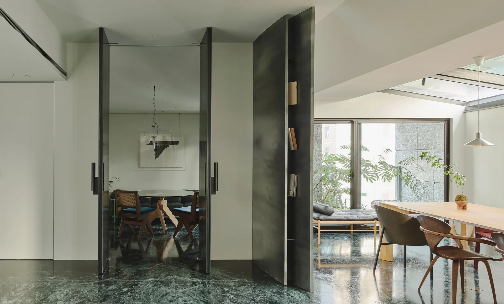 A modern dining room features a dark green marble floor, a charcoal metal room partition with built-in shelves, and warm wood mid-century chairs.