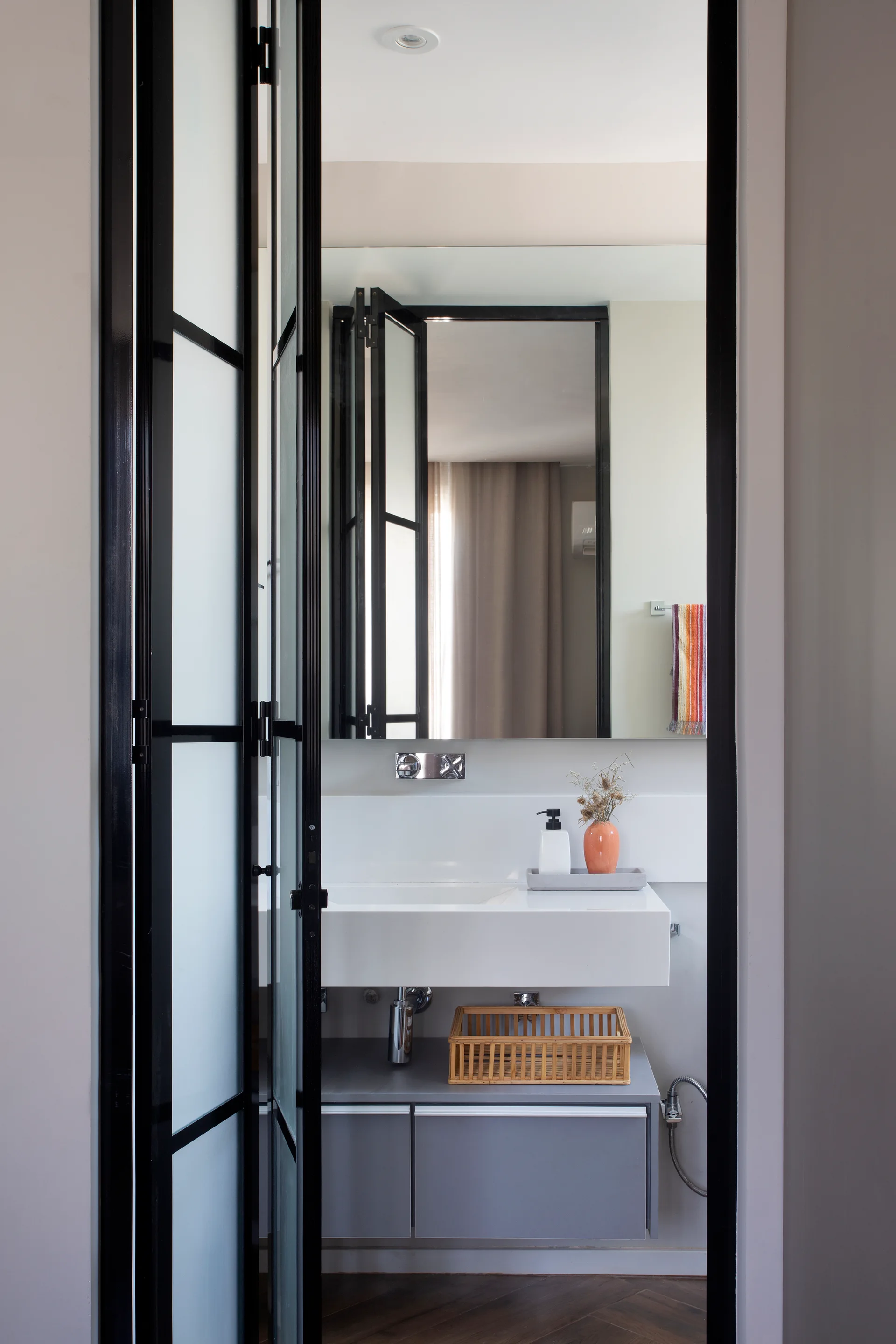 A modern bathroom viewed through a black-framed glass door, featuring a white floating vanity, a wall-mounted chrome faucet, and a grey cabinet.