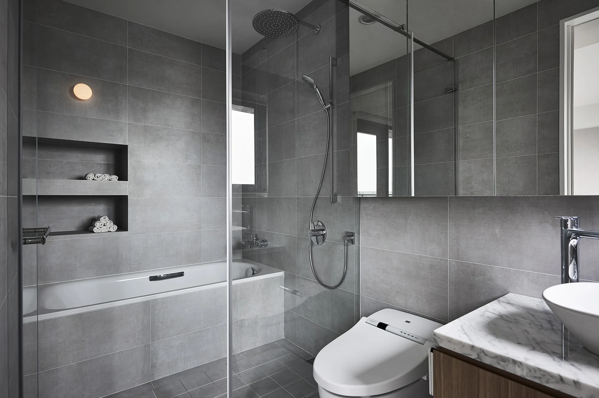 A modern bathroom featuring large grey wall tiles, a glass shower partition, a built-in bathtub, a white toilet, and a marble vanity counter.