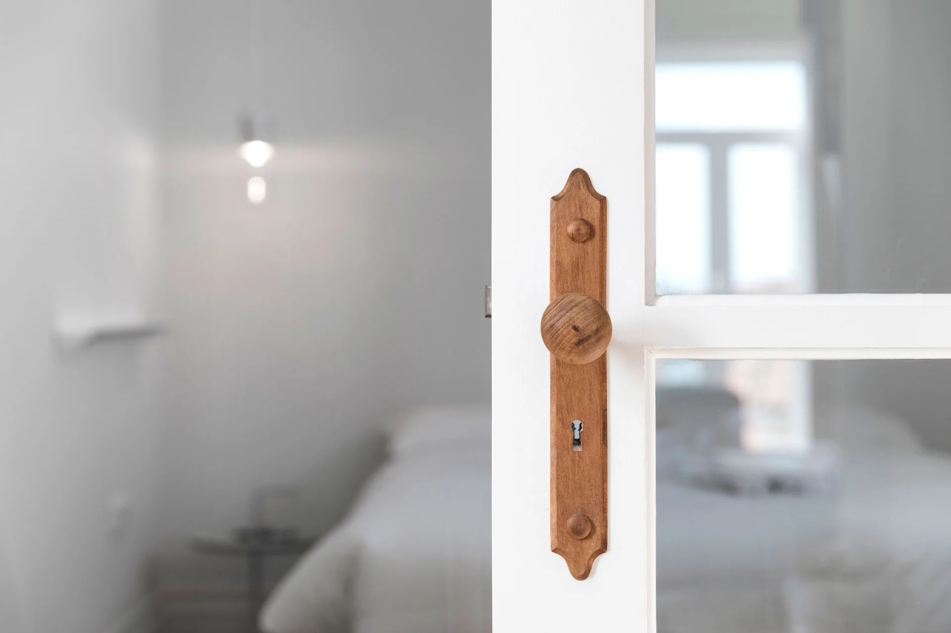 A close-up of a natural wood door knob and keyhole backplate on a white painted glass-paneled door opening into a blurred bedroom.