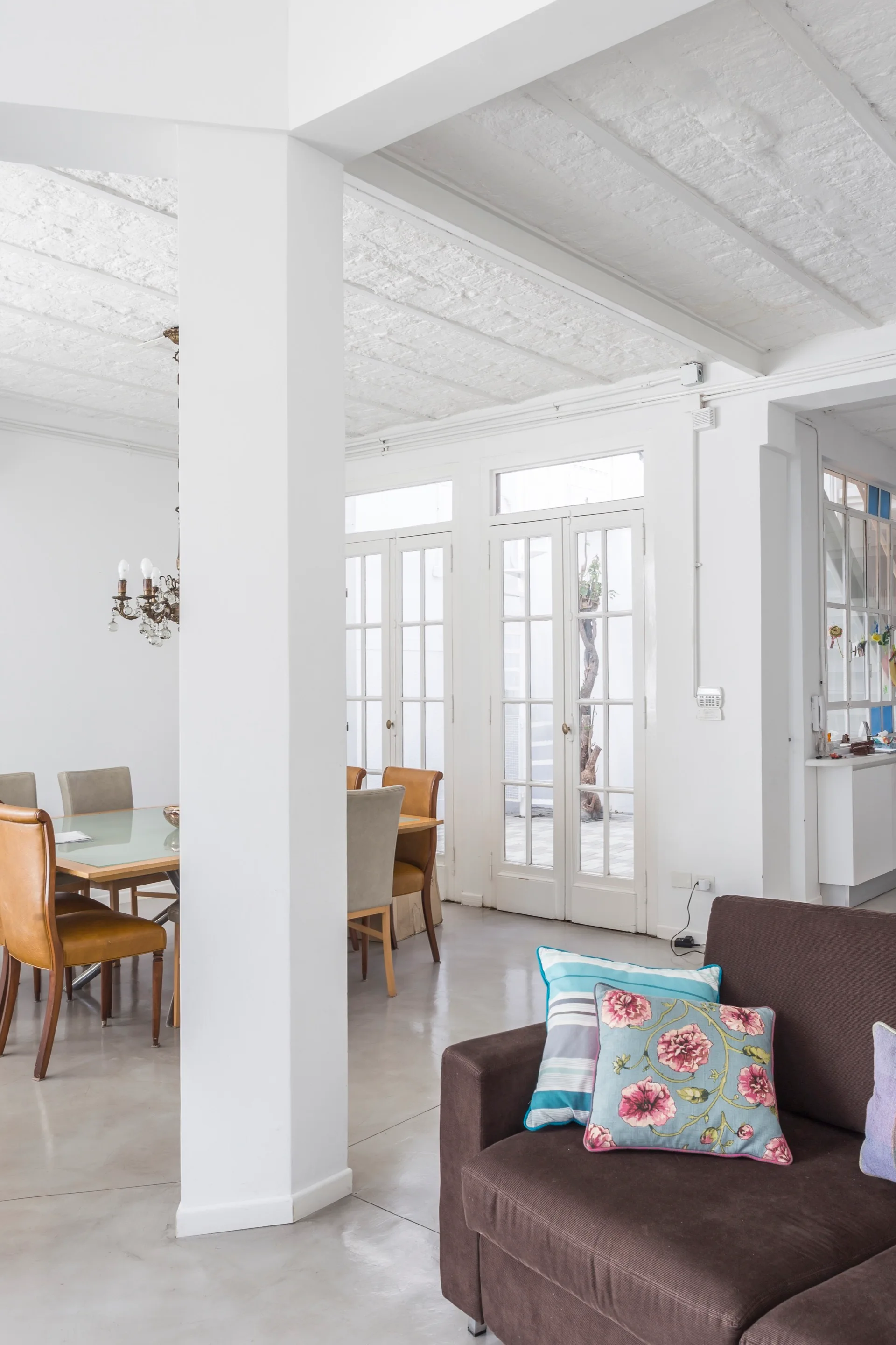 A bright room with a white structural column, a brown sofa with floral pillows, and a dining table set near multi-pane French doors.
