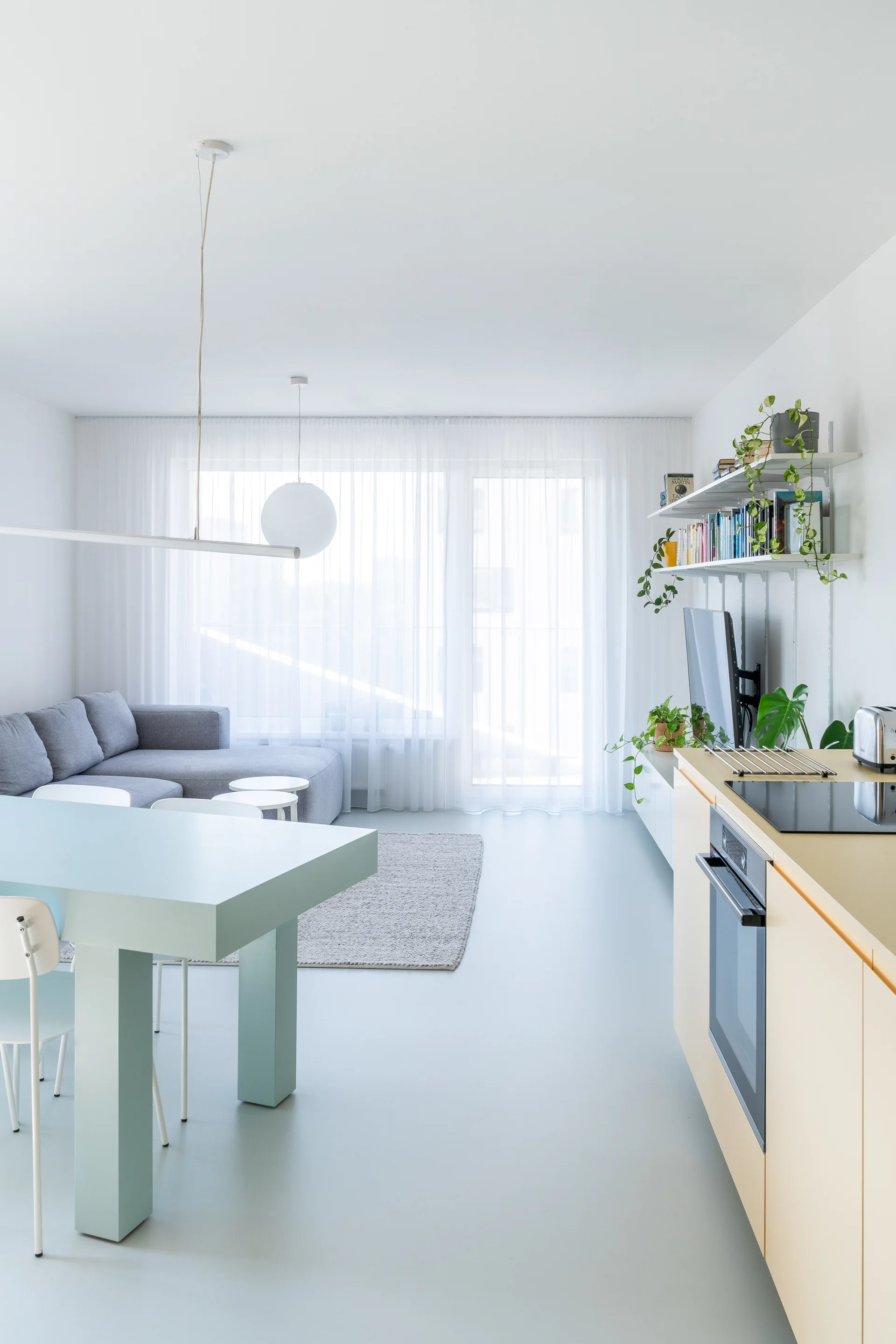A modern open-plan studio apartment featuring a mint blue dining table, grey sectional sofa, and light cream kitchen cabinets.