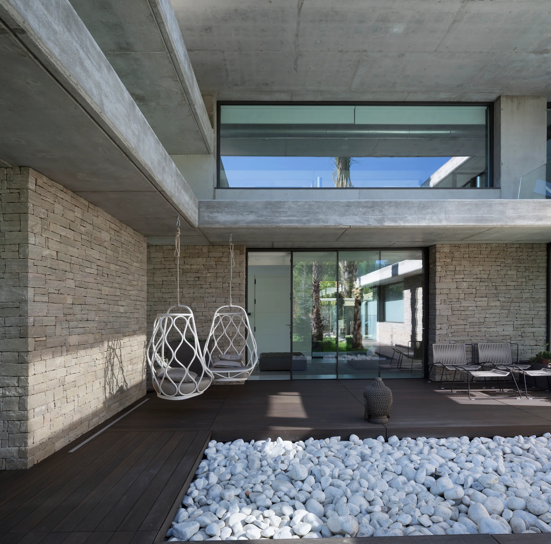 A modern patio features a dark wood deck with a white pebble bed, two hanging swing chairs, and textured stone walls.