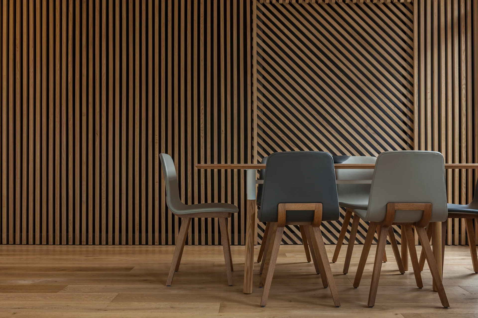 A wooden dining table and grey upholstered chairs are set against a wall made of vertical and diagonal light wood slats.