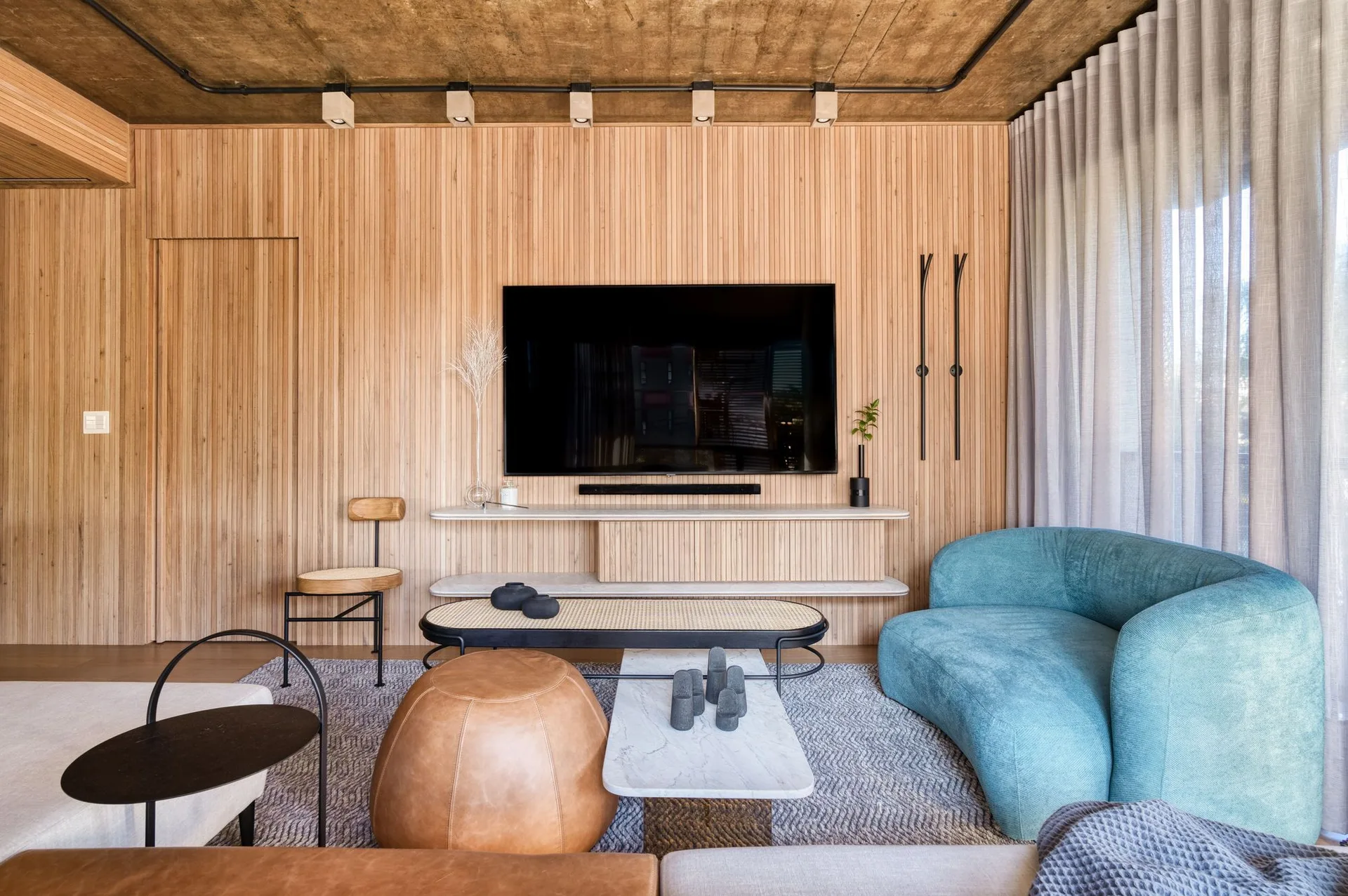 A contemporary living room featuring a vertical slatted wood wall, an exposed concrete ceiling, a television, and a curved blue sofa on a grey rug.