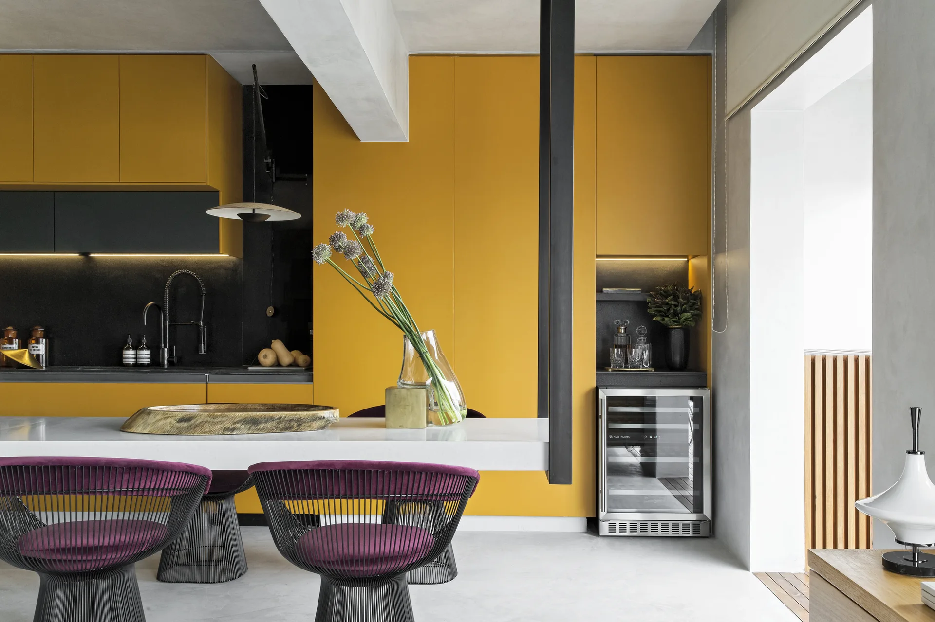 A modern kitchen features full-height mustard yellow cabinets, a white stone island with purple velvet chairs, and a built-in wine cooler.