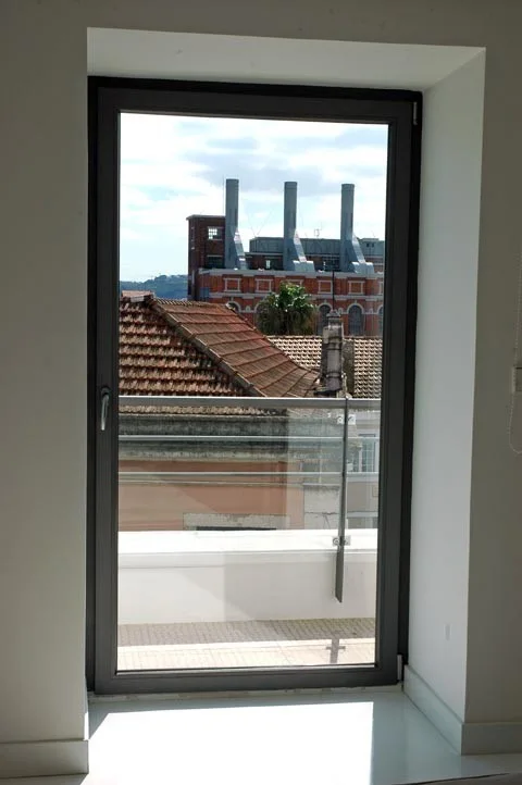 A large rectangular dark grey metal window set deep in a white wall looks out onto terracotta roofs and brick city buildings.