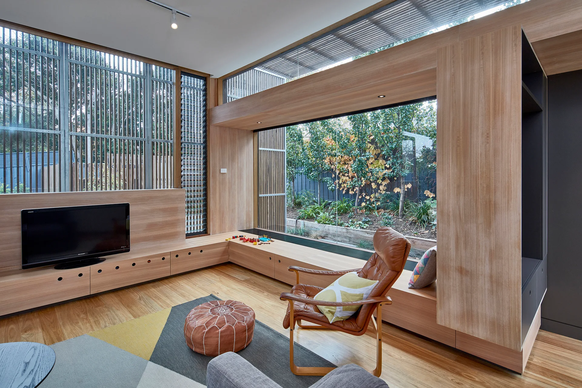 A contemporary living room featuring light wood built-in cabinetry, a low media unit, a tan leather armchair, and a large window looking onto a garden.