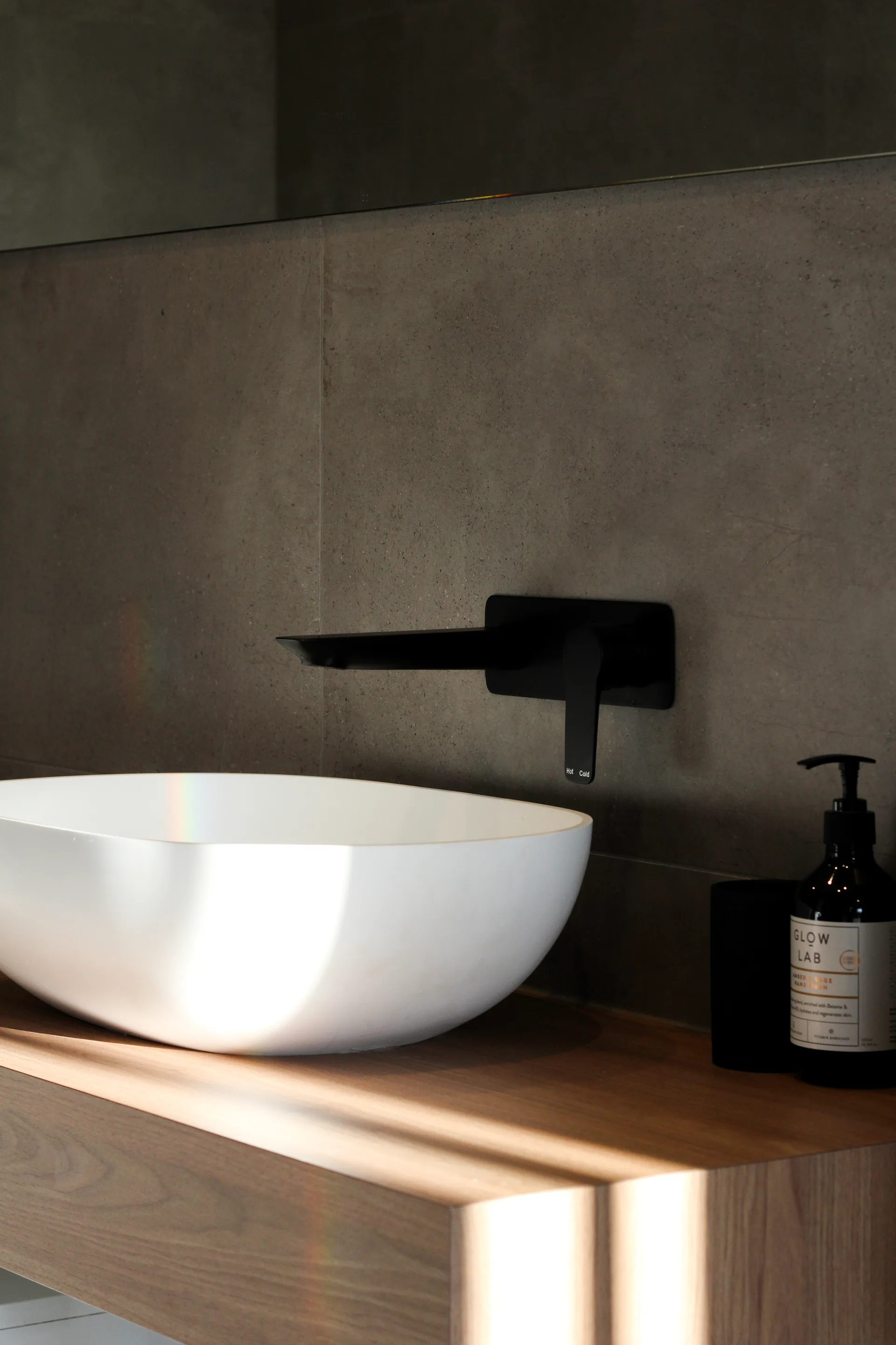 A minimalist bathroom vanity features a white oval vessel sink on a wood countertop with a wall-mounted matte black faucet against grey concrete tiles.