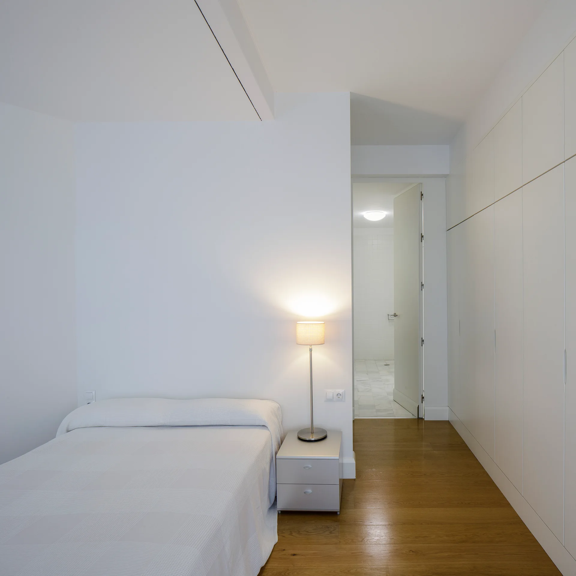 A minimalist white bedroom featuring a low bed, a metallic nightstand with an illuminated lamp, and built-in floor-to-ceiling closets.