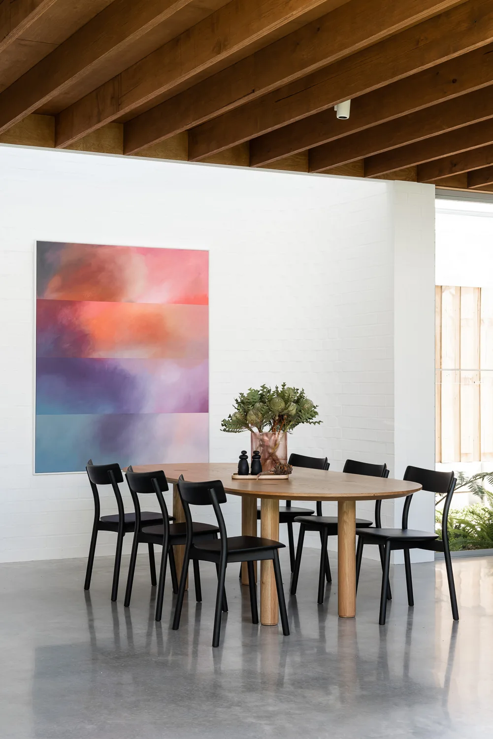A modern dining room features an oval wooden table surrounded by black chairs beneath an exposed wood ceiling and next to a large colorful painting.