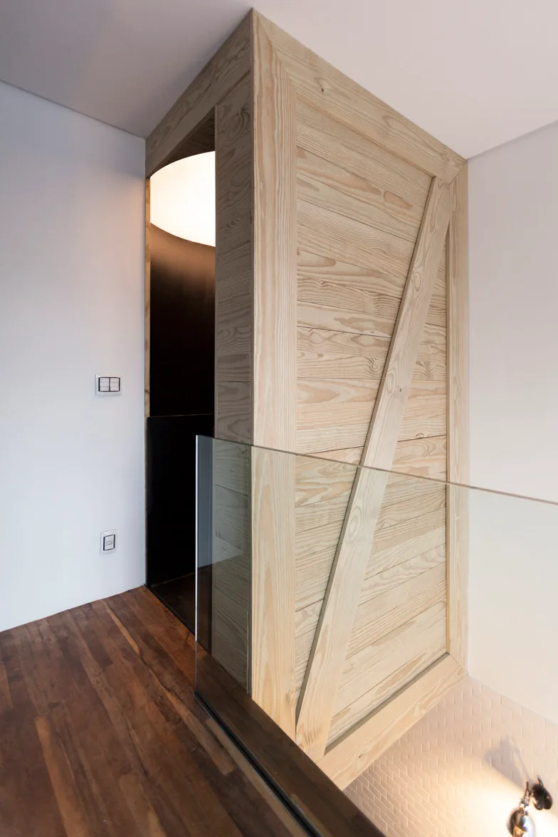 A modern staircase landing featuring a light-toned wooden panel wall partition with diagonal bracing next to a transparent glass railing and dark wood floors.