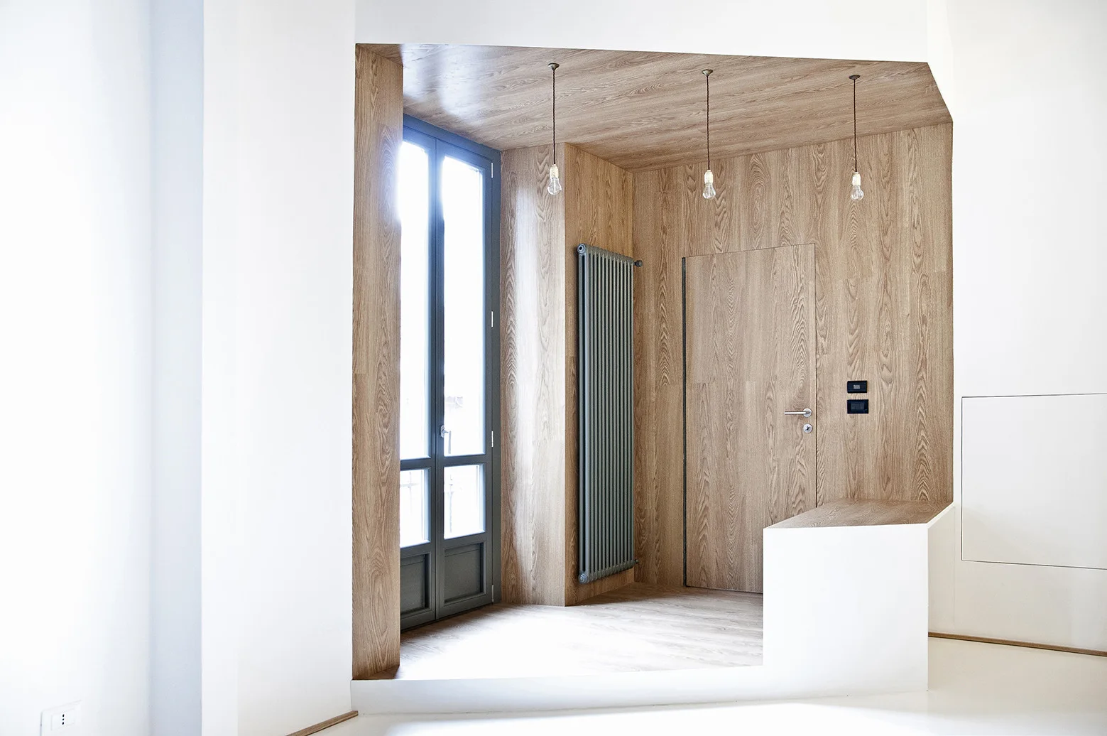 A modern entryway features a recessed area lined with warm wood paneling, a flush matching door, three bare bulb pendants, and a tall grey radiator.