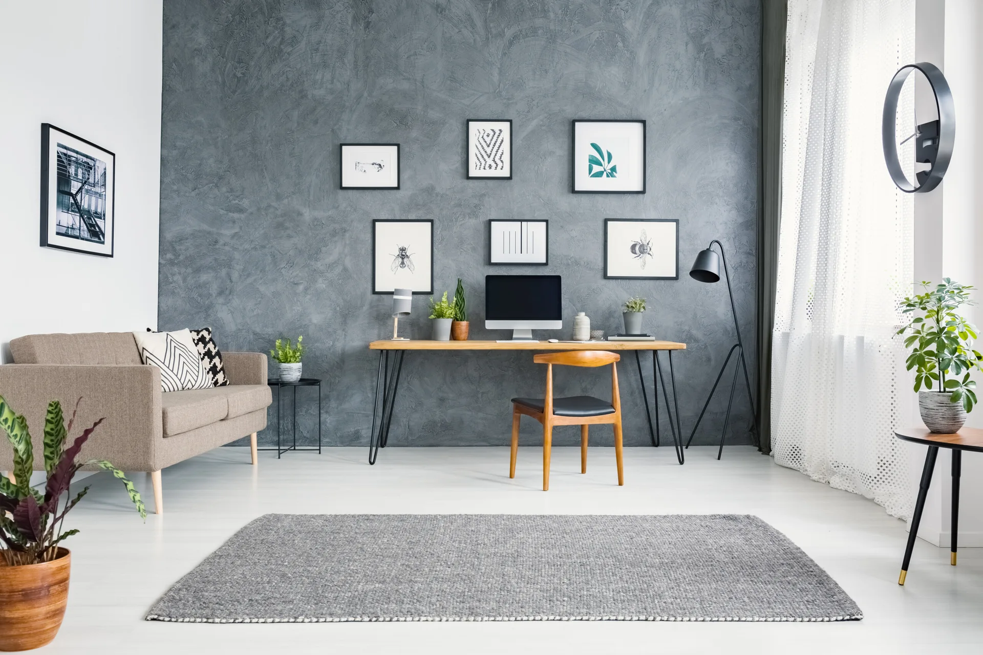 A light-filled home office features a wooden desk, mid-century chair, and black-framed art gallery on a textured gray plaster accent wall.