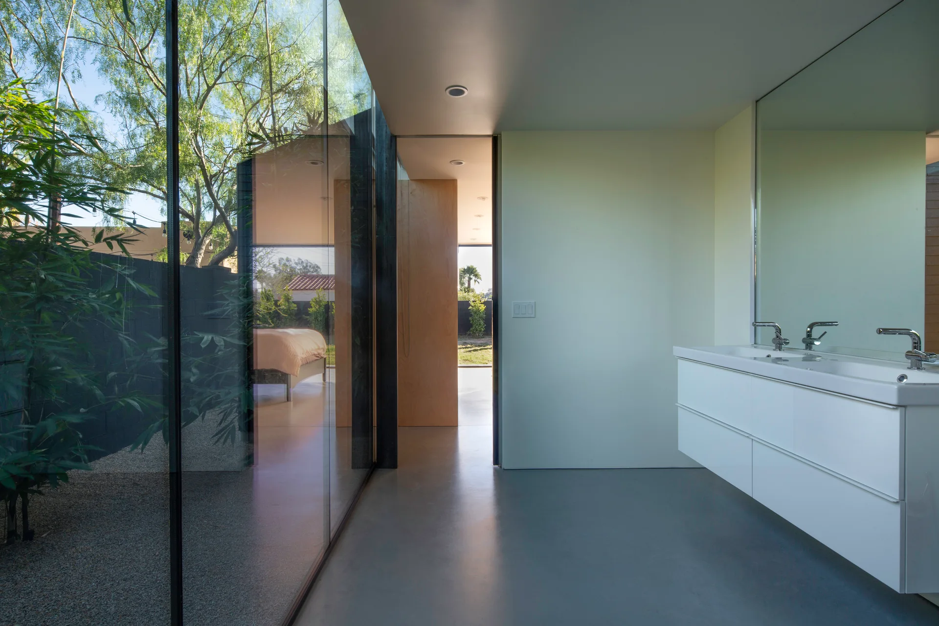 A modern minimalist bathroom features a wall-mounted white vanity, a frameless mirror, and a large floor-to-ceiling glass wall overlooking a courtyard.