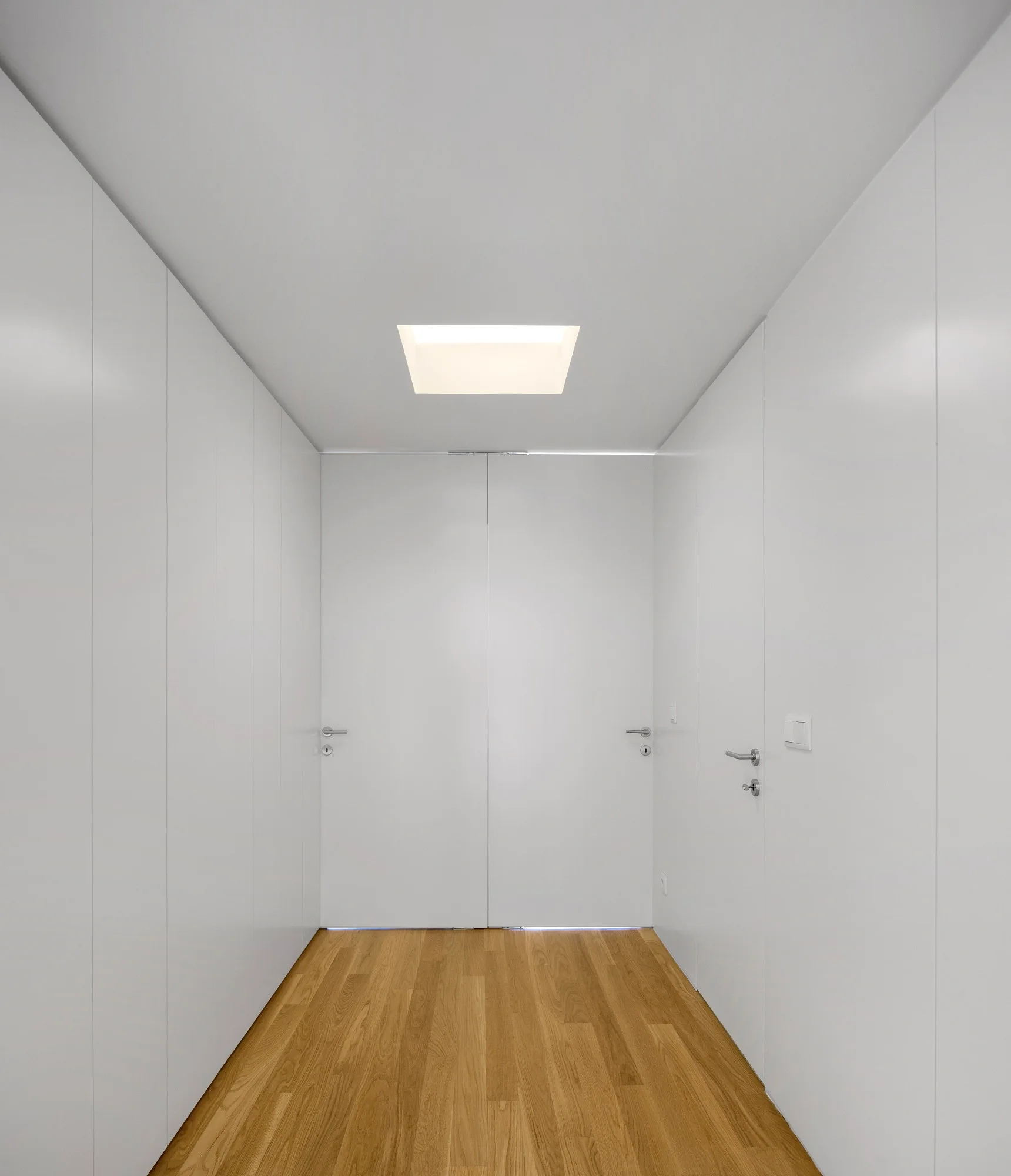 A minimalist white hallway features integrated flush doors, a light oak plank floor, and a square skylight in the ceiling.