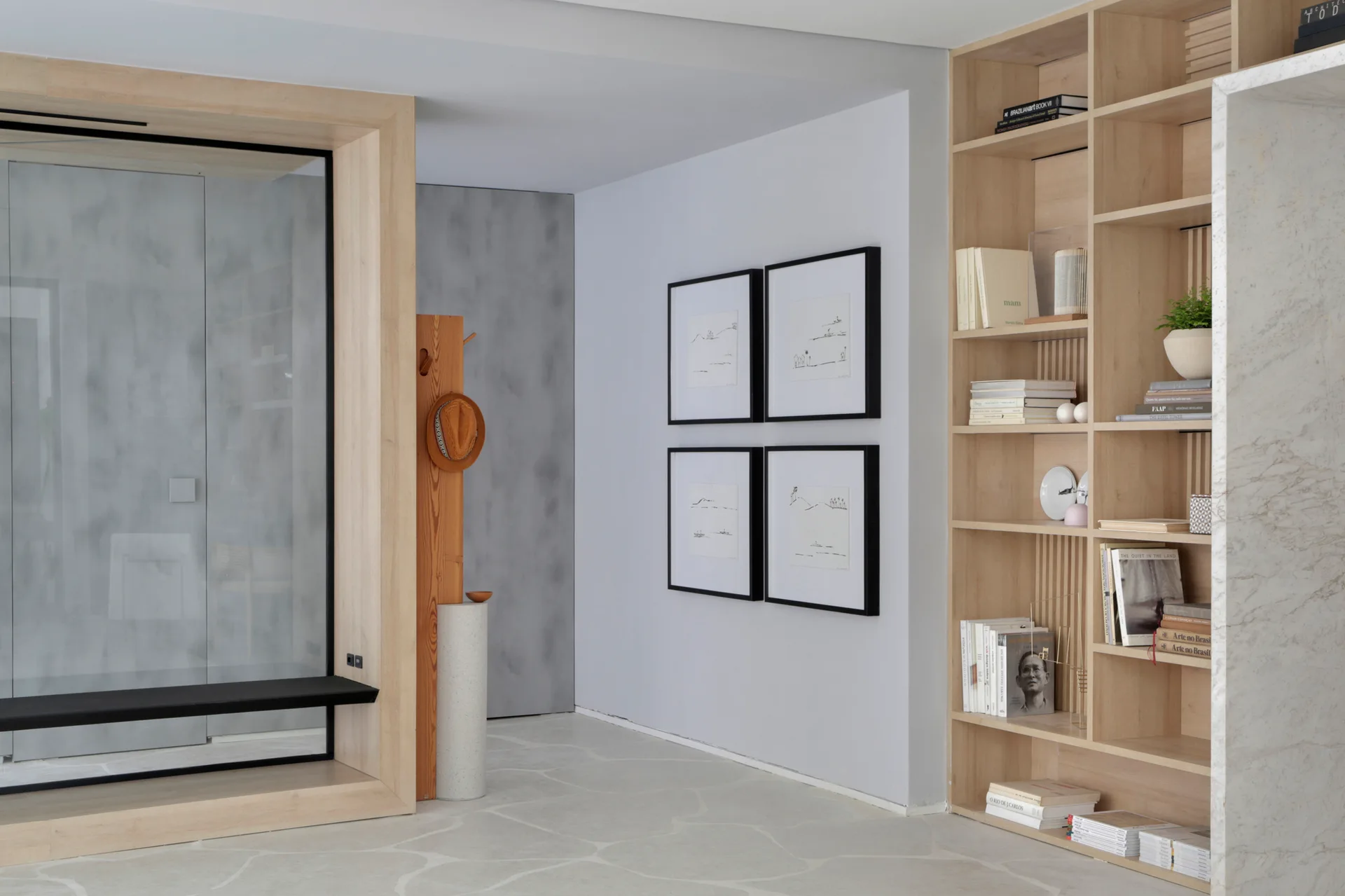 A modern entryway with a light wood custom bookshelf, a black bench framed in wood, and four framed drawings on the wall.