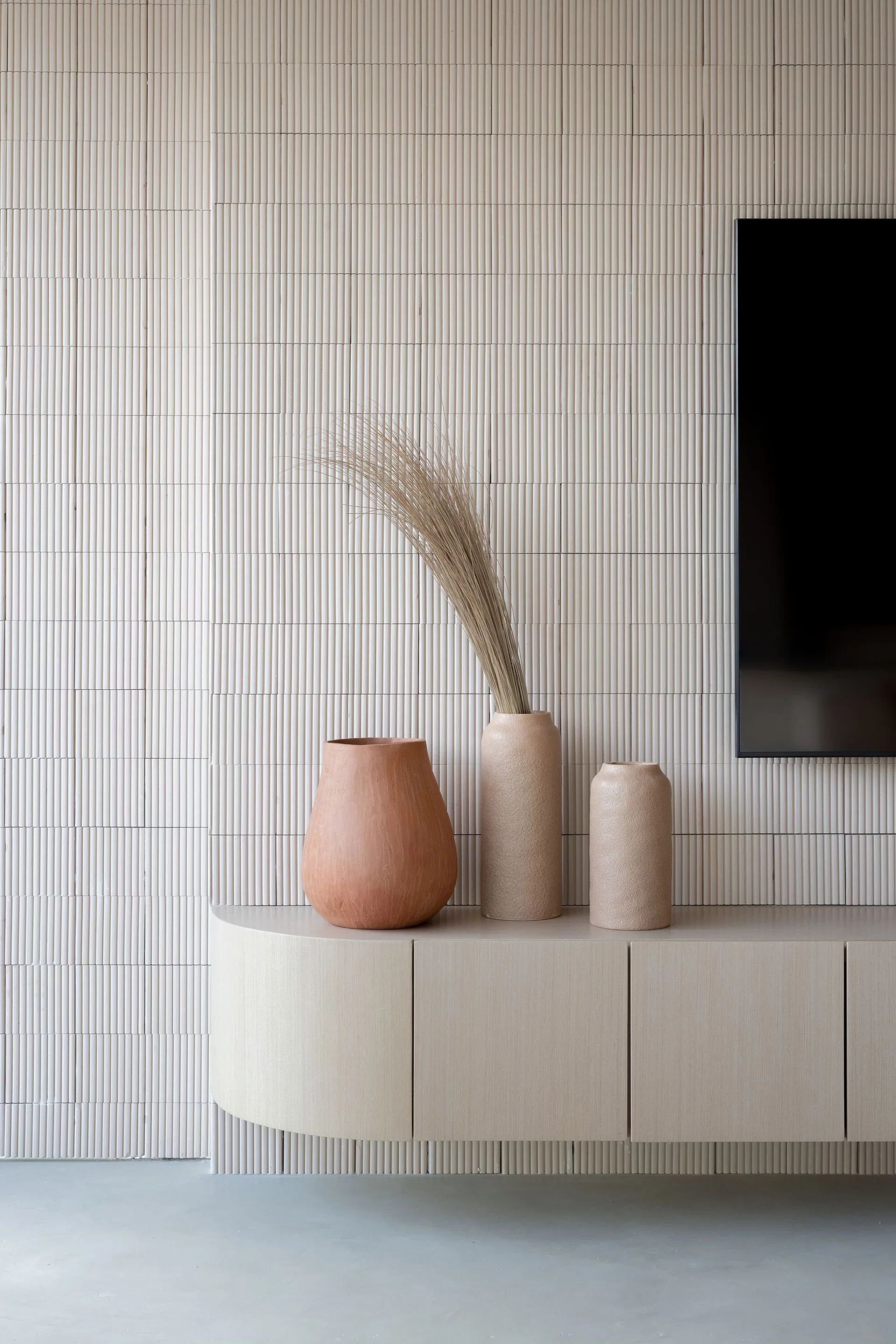 A beige floating console cabinet with a curved corner sits below a black TV screen against a vertically fluted wall with three ceramic vases.