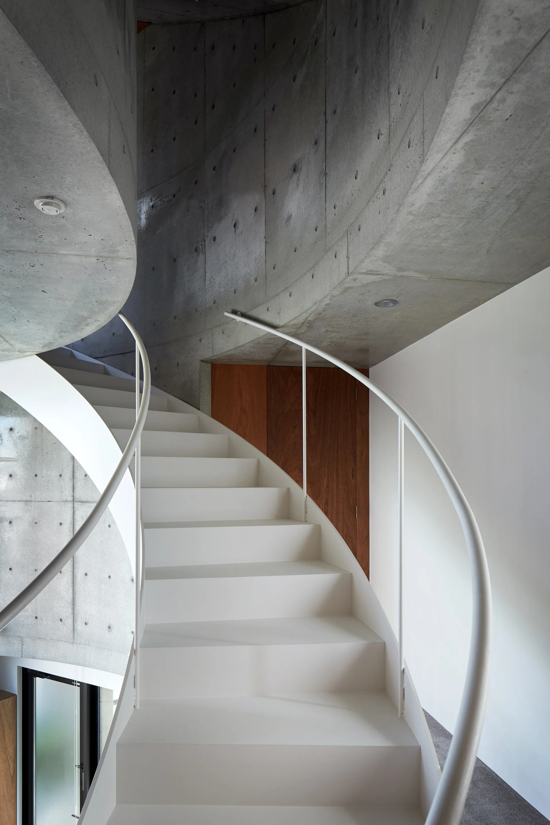 A curved white staircase with a slender metal handrail rises alongside a smooth, curved concrete wall with visible circular markings.