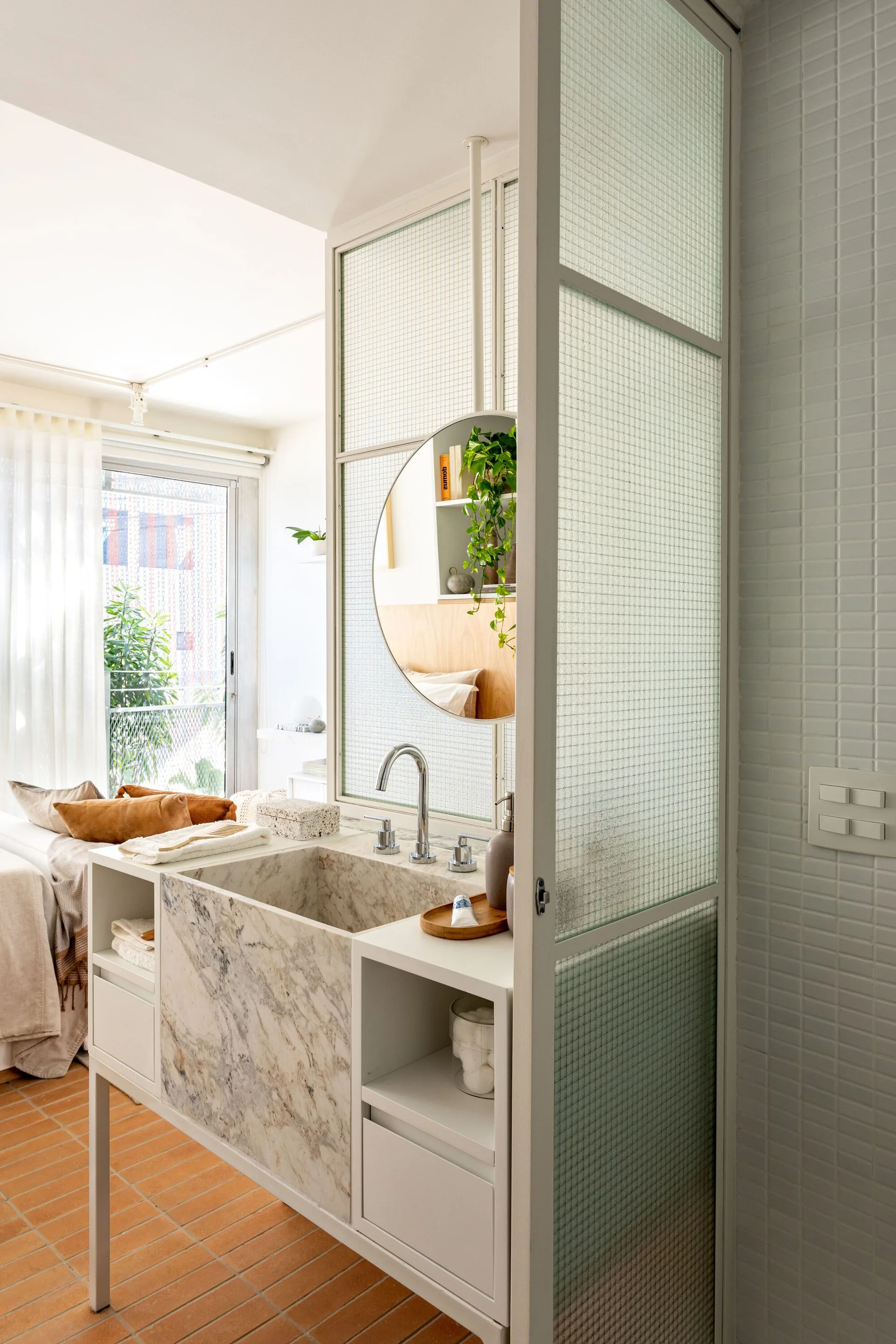 A white bathroom vanity with a marble sink sits beside a gridded textured glass partition screen, with terracotta floor tiles below.