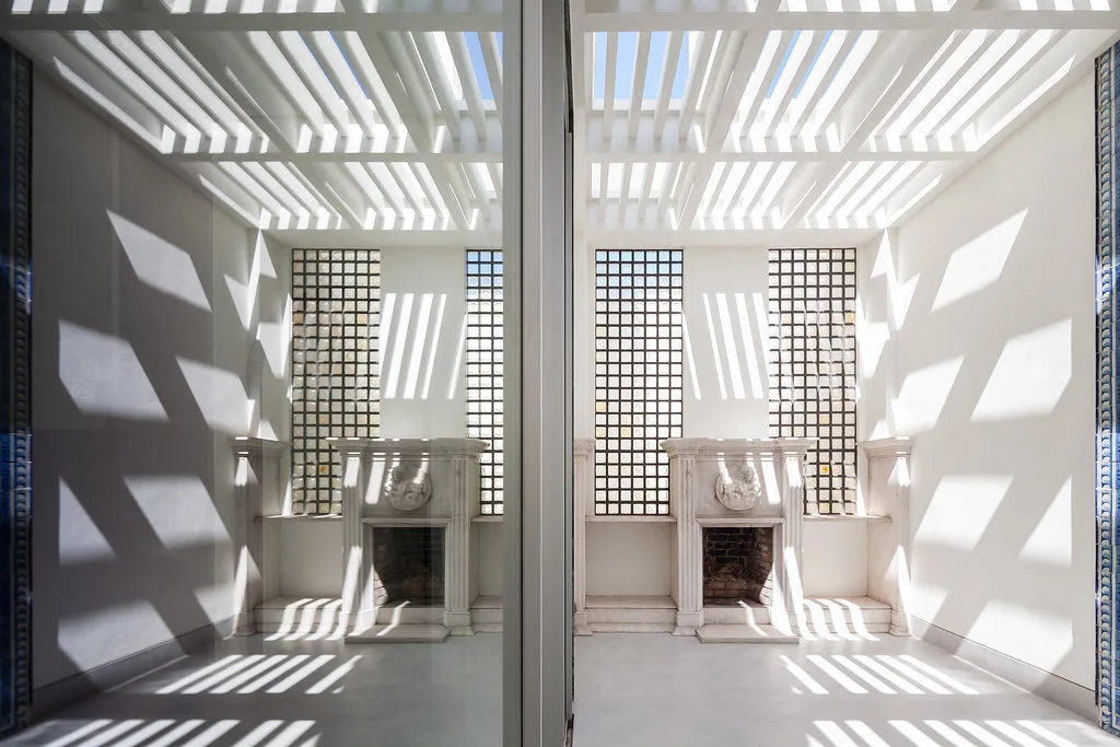 A bright white sunroom features a classical carved stone fireplace beneath a slatted ceiling that casts bold, striped shadows across the room.