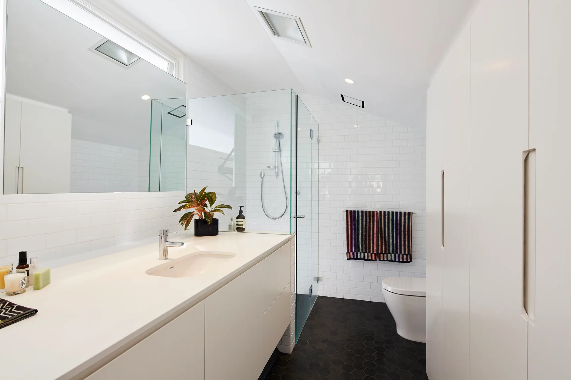 A modern white bathroom with subway tiled walls, dark hexagonal floor tiles, a glass shower enclosure, and a minimalist vanity sink.