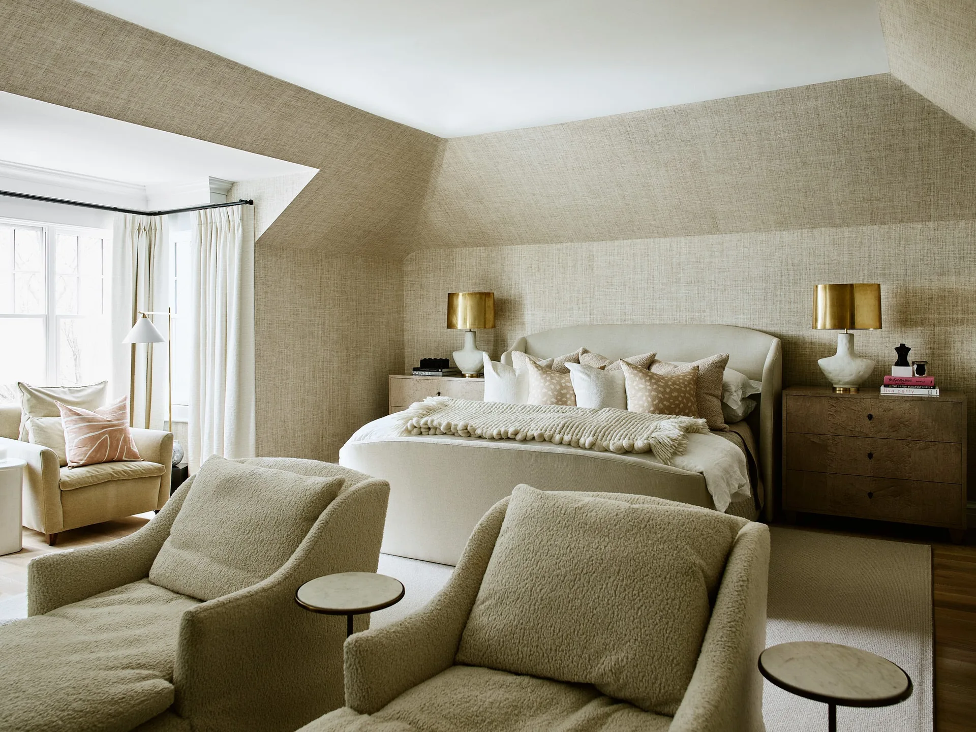A neutral beige bedroom featuring a bed flanked by wooden nightstands with brass lamps, under a sloped ceiling wrapped in textured wallpaper.