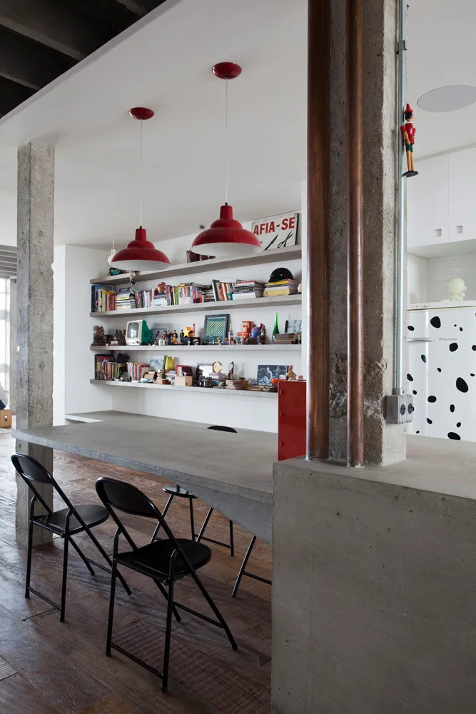 An industrial dining space featuring a long concrete dining table, black folding chairs, red pendant lights, and concrete floating wall shelves.