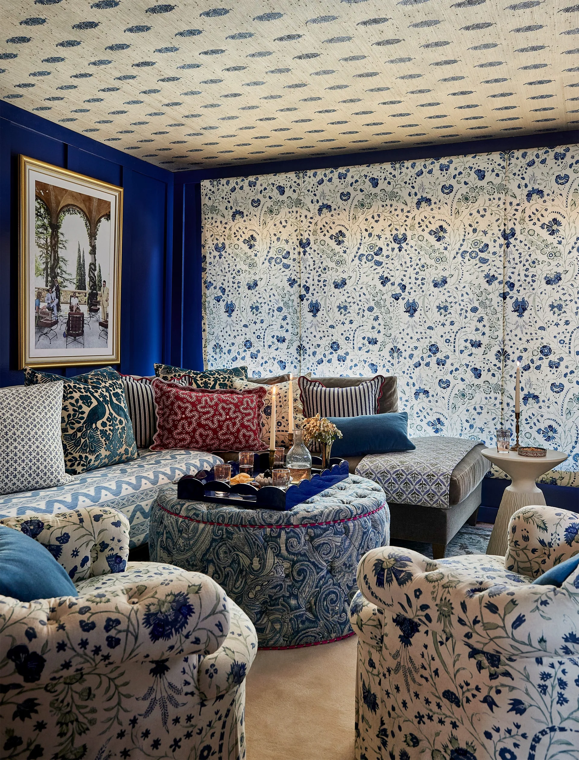 A blue and white maximalist living room features patterned wallpaper on the walls and ceiling, floral armchairs, and a large tufted paisley ottoman.