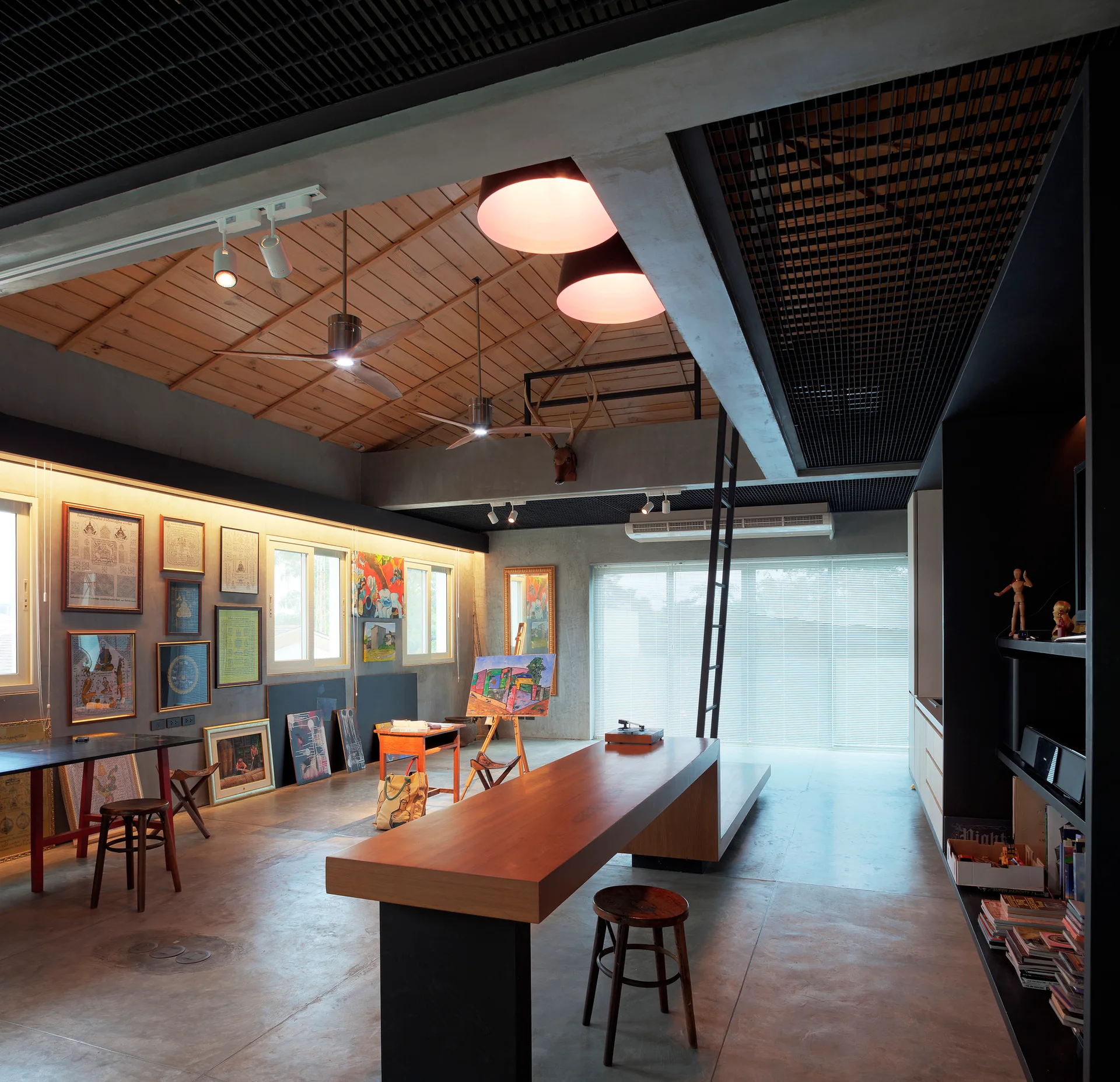 An industrial creative studio features concrete floors, a sloped wood ceiling, a long timber work table, and framed artwork resting along the wall.