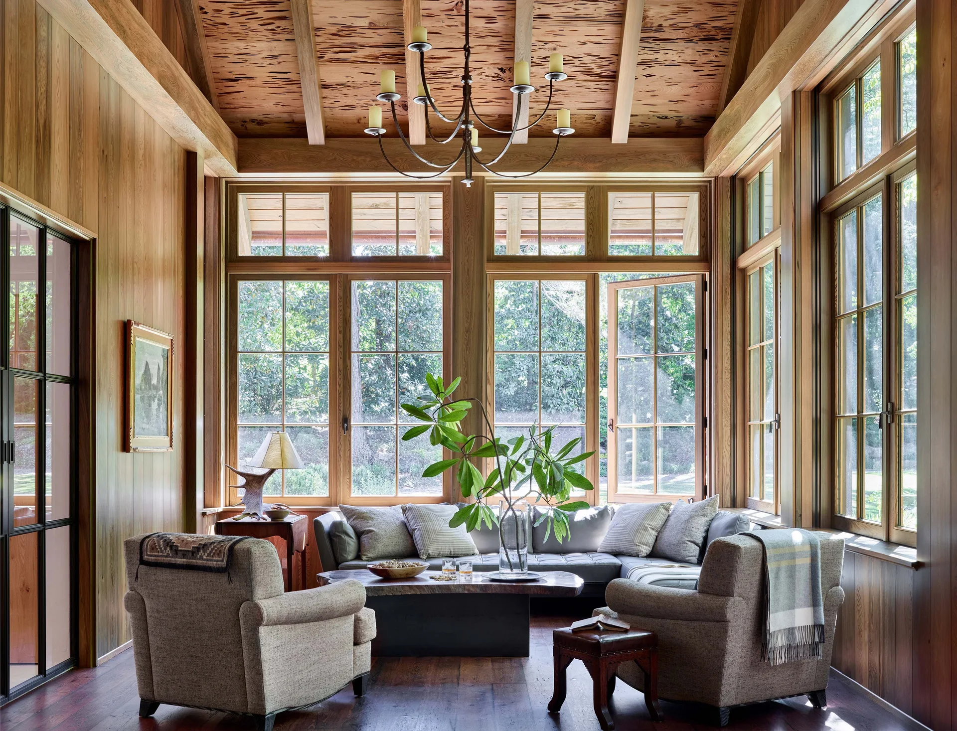 A rustic living room with wood-paneled walls, a vaulted ceiling, large multi-pane windows, a grey sectional sofa, and two upholstered armchairs.