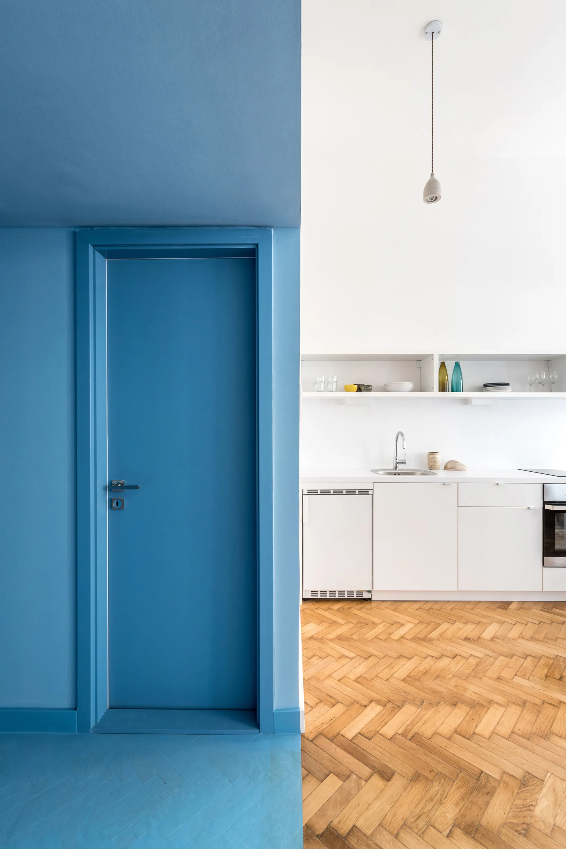 A minimalist white kitchen with herringbone wood floors sits adjacent to a solid blue painted entryway with a matching blue door.