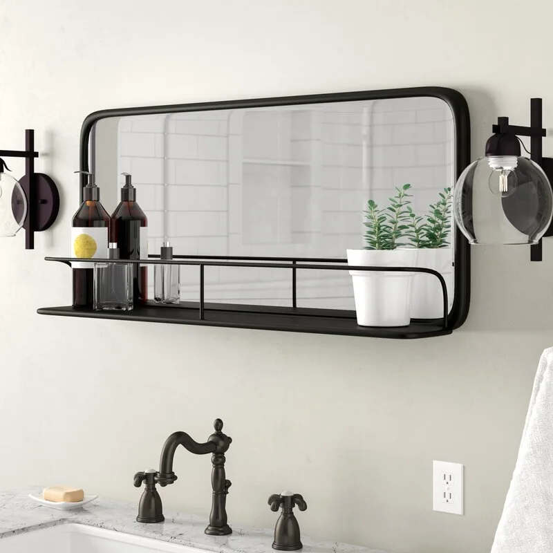 A rectangular matte black bathroom mirror with an integrated metal storage shelf is mounted above a marble vanity with globe wall sconces.