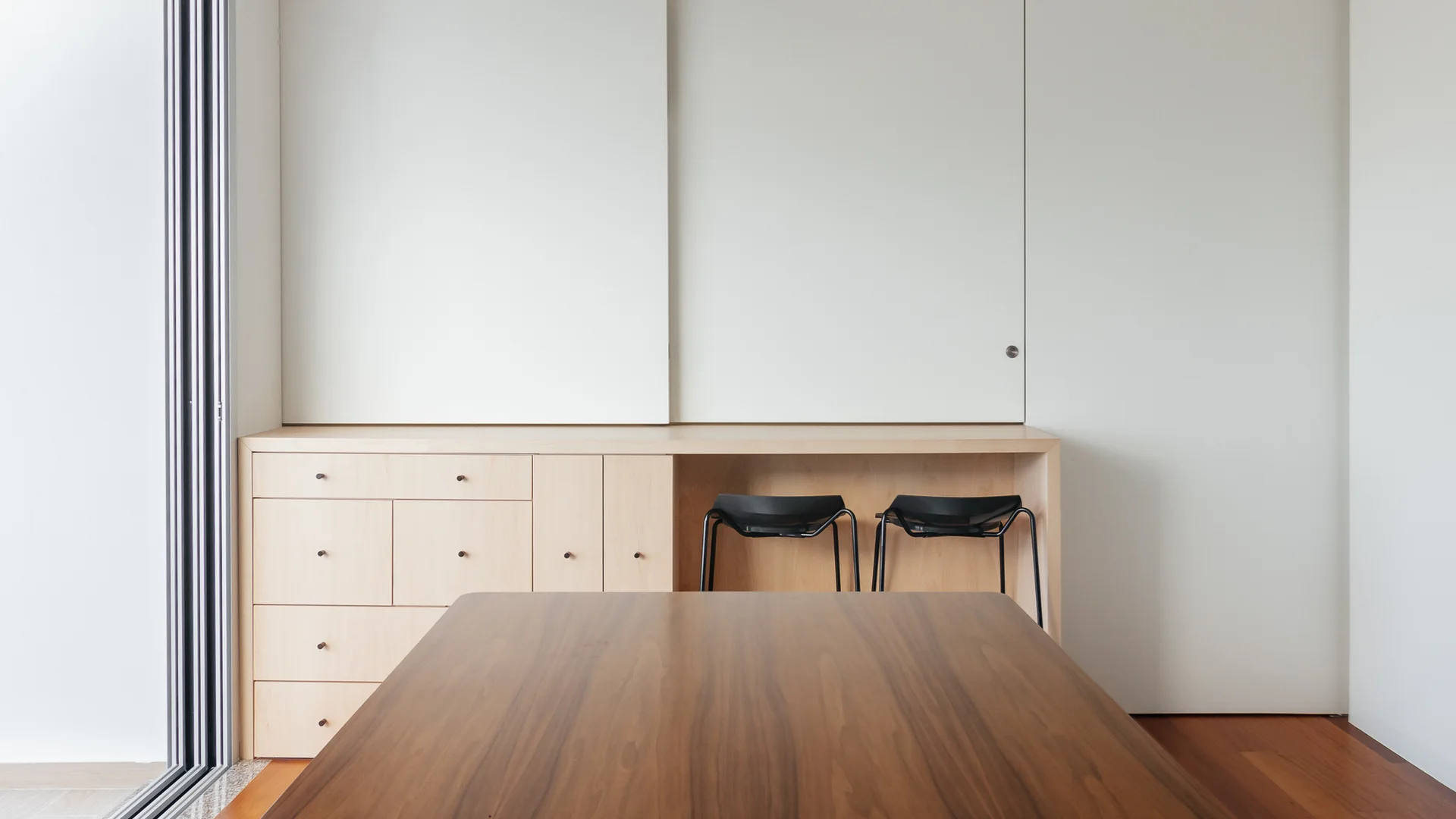 A modern dining room featuring a dark wood table, light wood built-in cabinets with two tucked-away black stools, and white sliding wall panels.