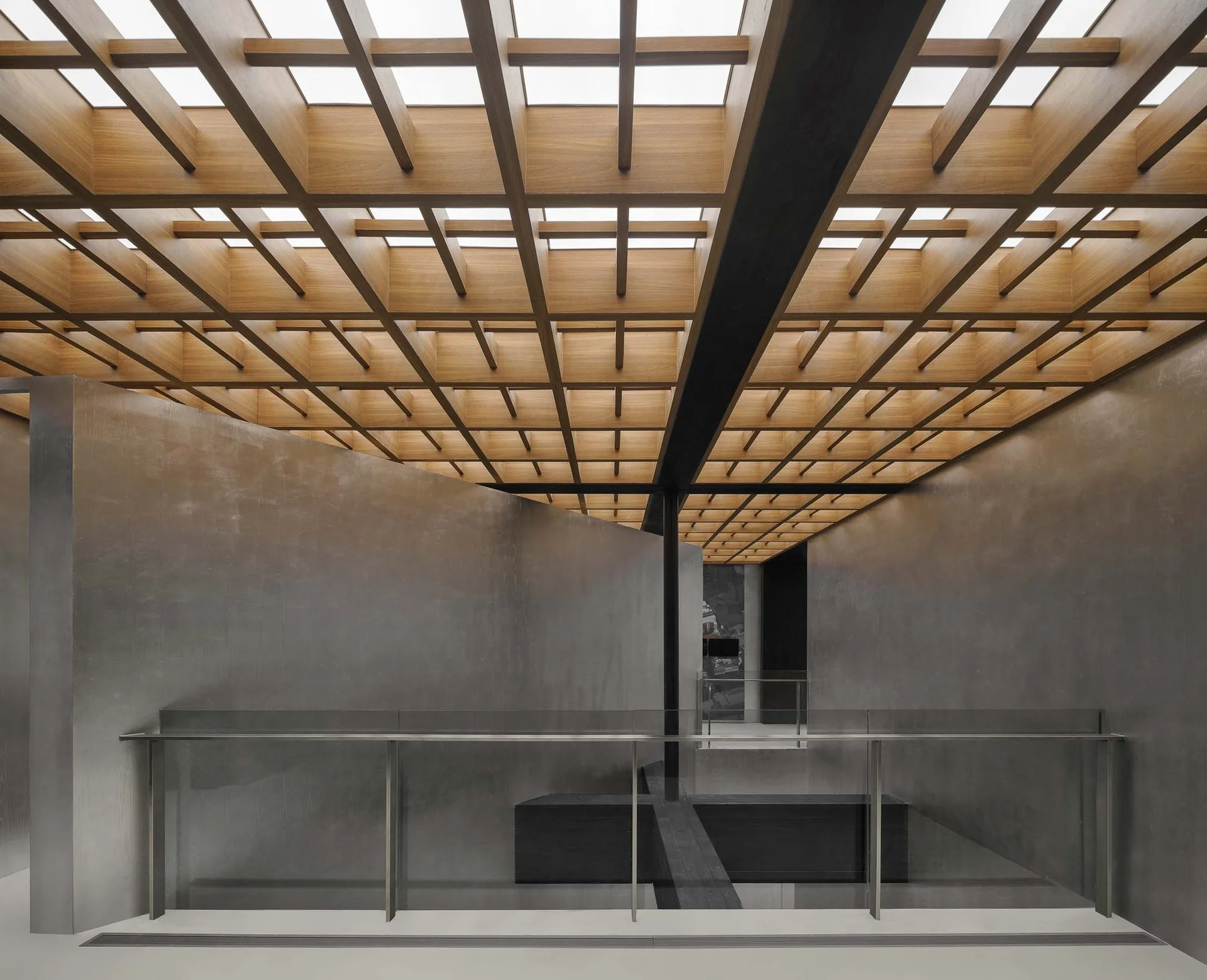 A modern staircase landing features brushed grey metal wall panels, a glass balustrade, and a large wooden grid ceiling with skylights.