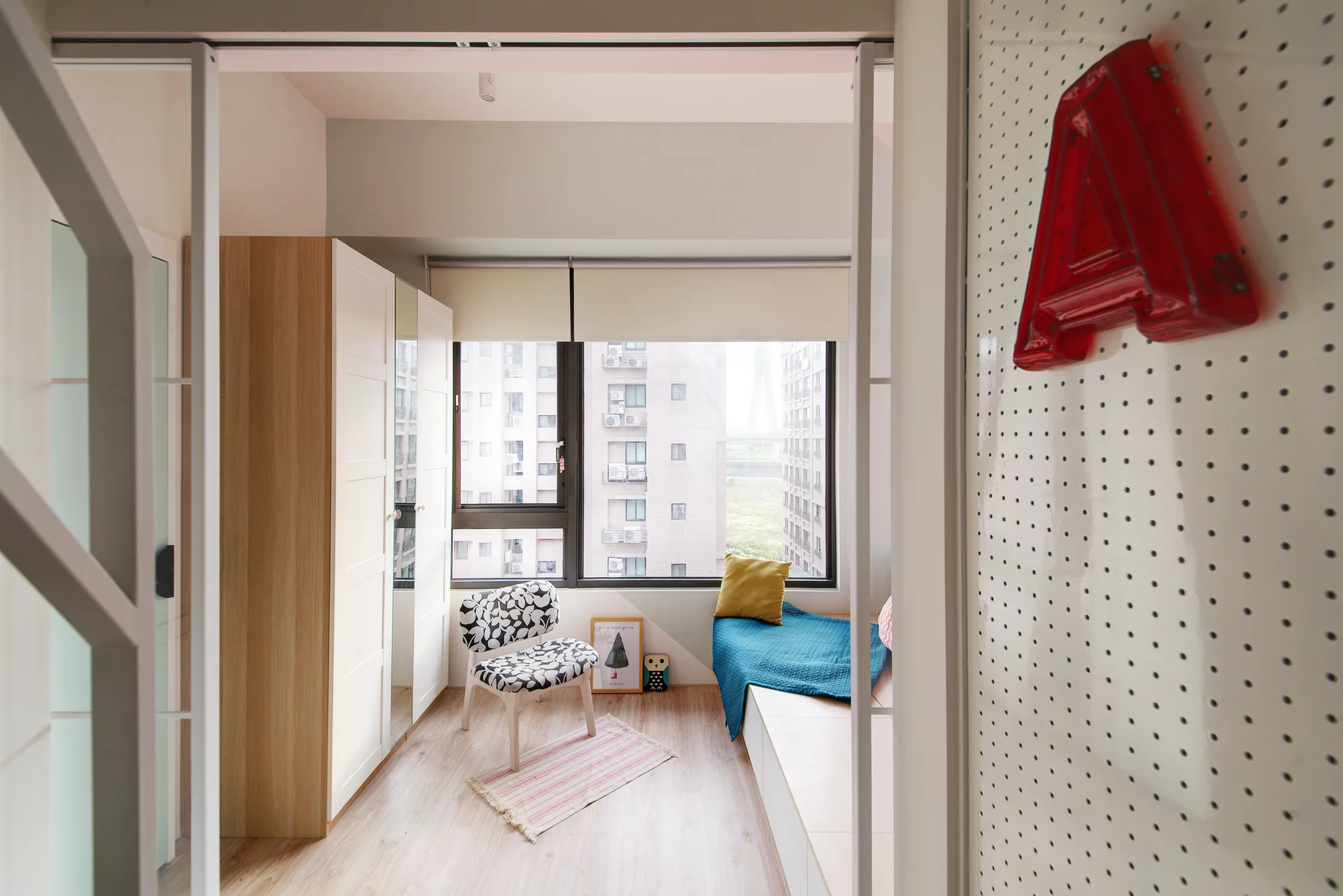 A small bedroom featuring a built-in wooden window bench, a black and white patterned accent chair, and a white pegboard wall.
