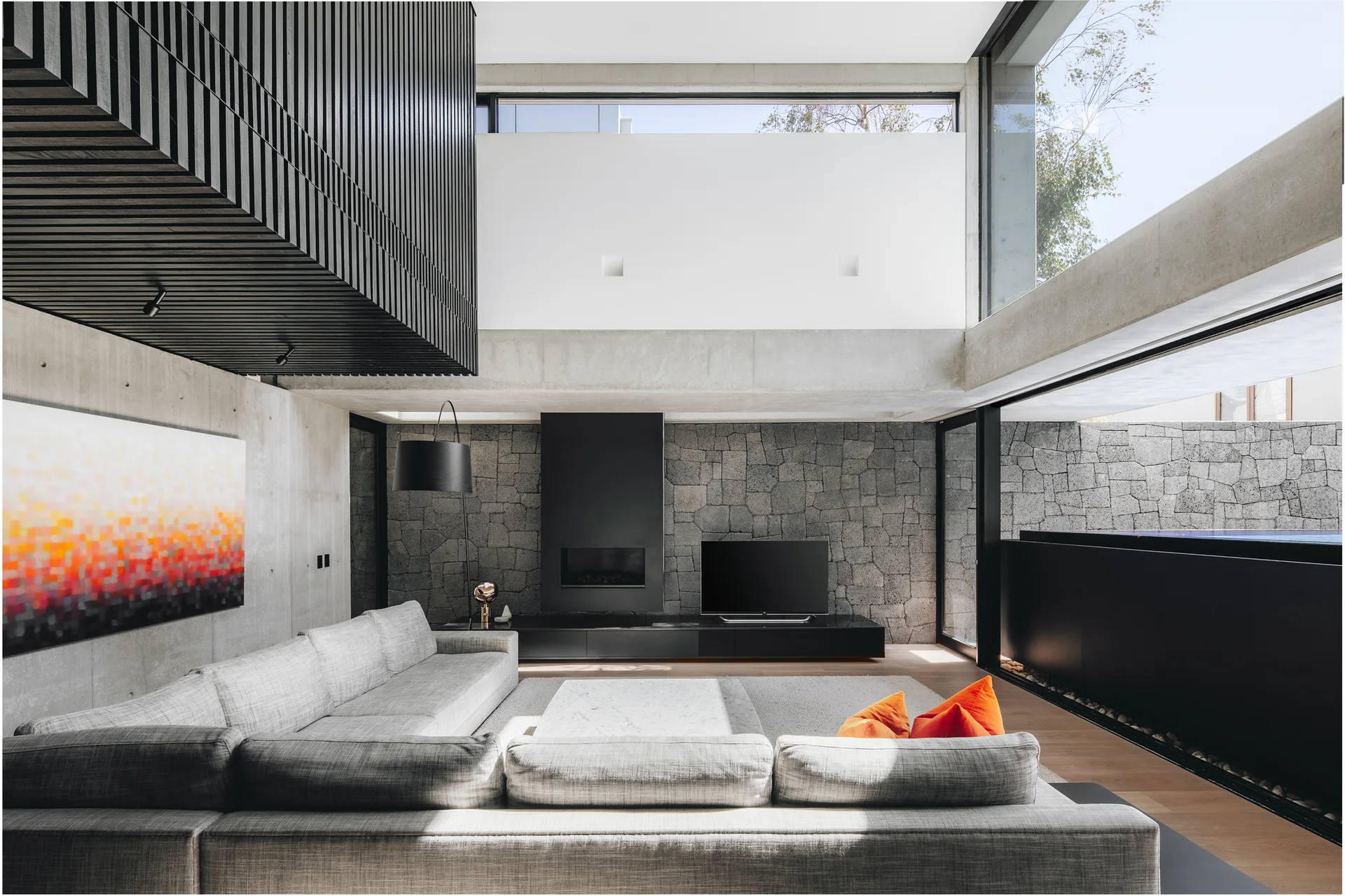 A modern living room featuring a large grey sectional sofa, exposed concrete walls, a textured stone fireplace wall, and a black wood slatted ceiling.