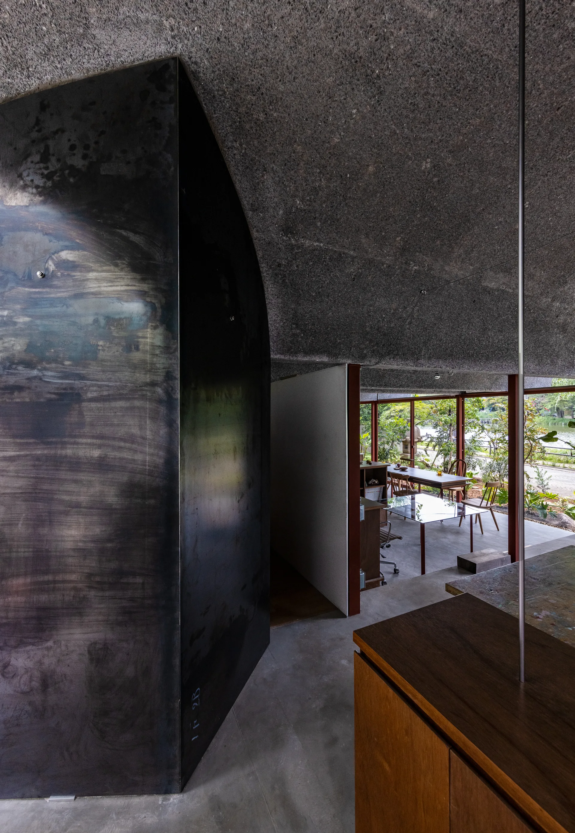 A modern minimalist interior featuring a curved textured concrete ceiling, a brushed metal wall, and a dining table visible in the background.